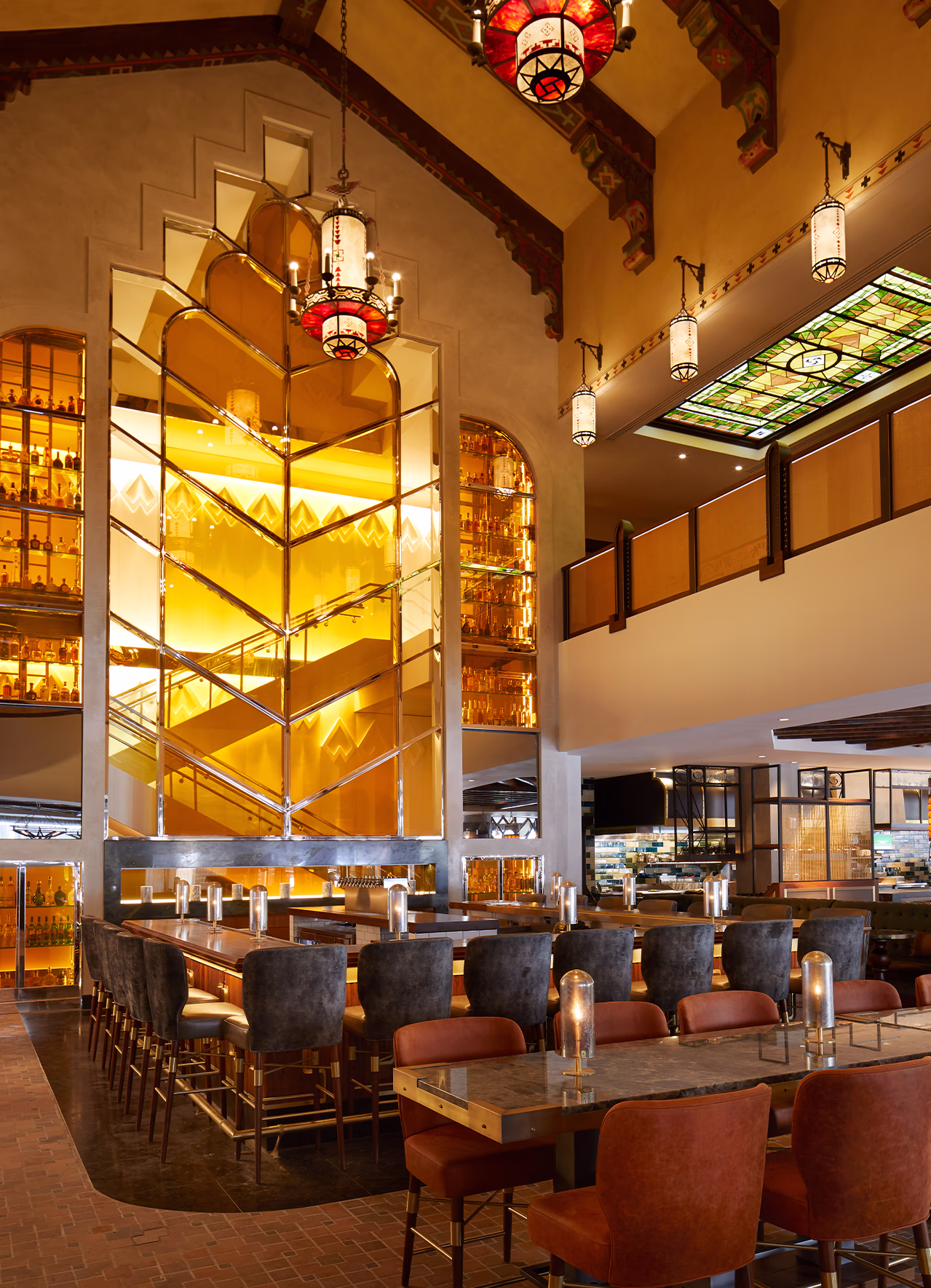 Double-height atrium at Ámbar Restaurante with a concealed staircase behind the amber glass tequila wall, beneath a Spanish Colonial beamed ceiling.