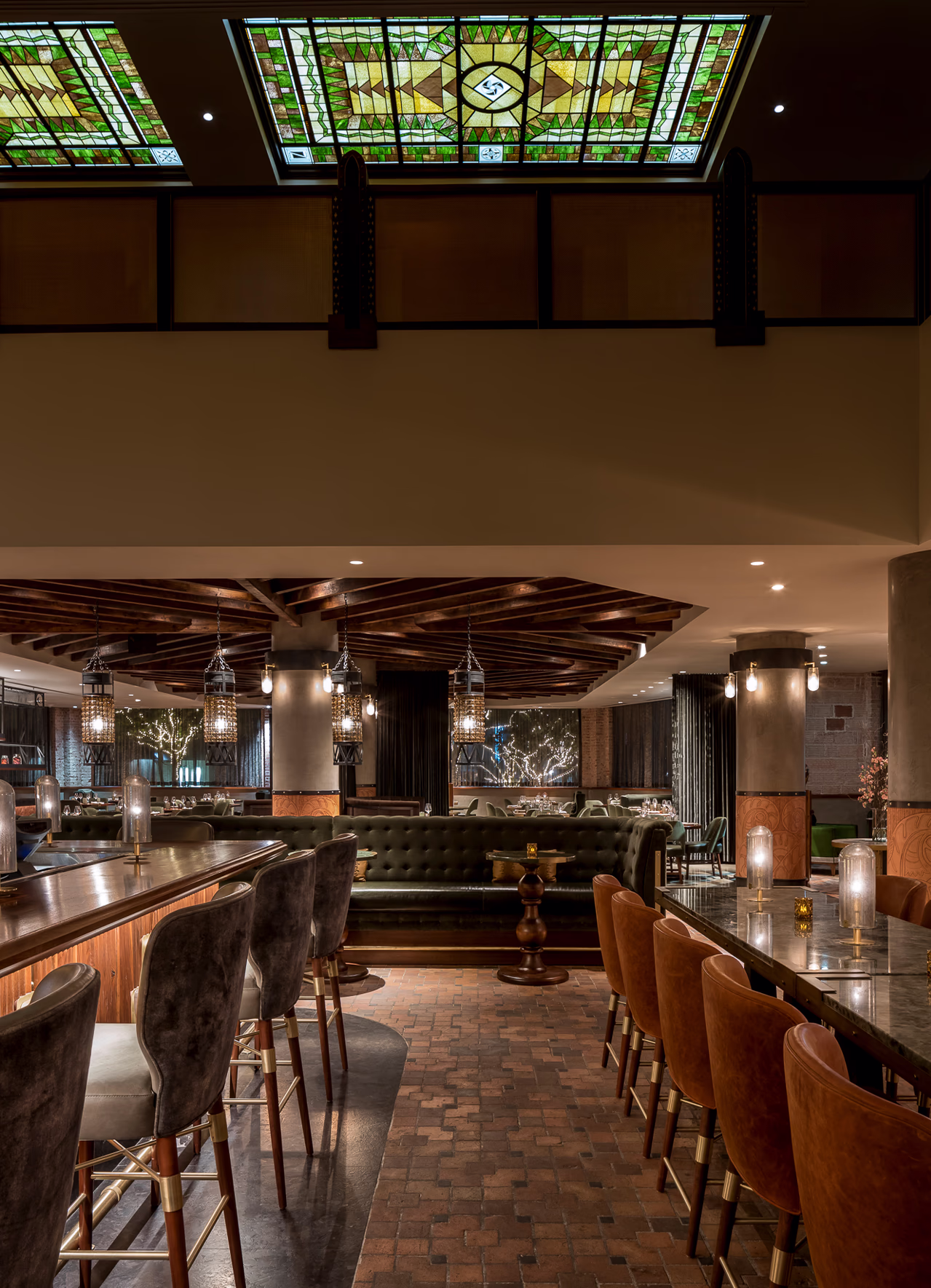 Bar seating with velvet and leather stools beneath original stained-glass skylights at Ámbar Restaurante, overlooking the dining room and lit trees.