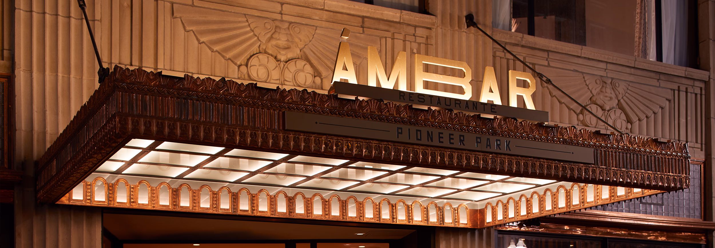Ornate copper art deco canopy with backlit Ámbar Restaurante signage marking the entrance to the historic Plaza Hotel Pioneer Park.