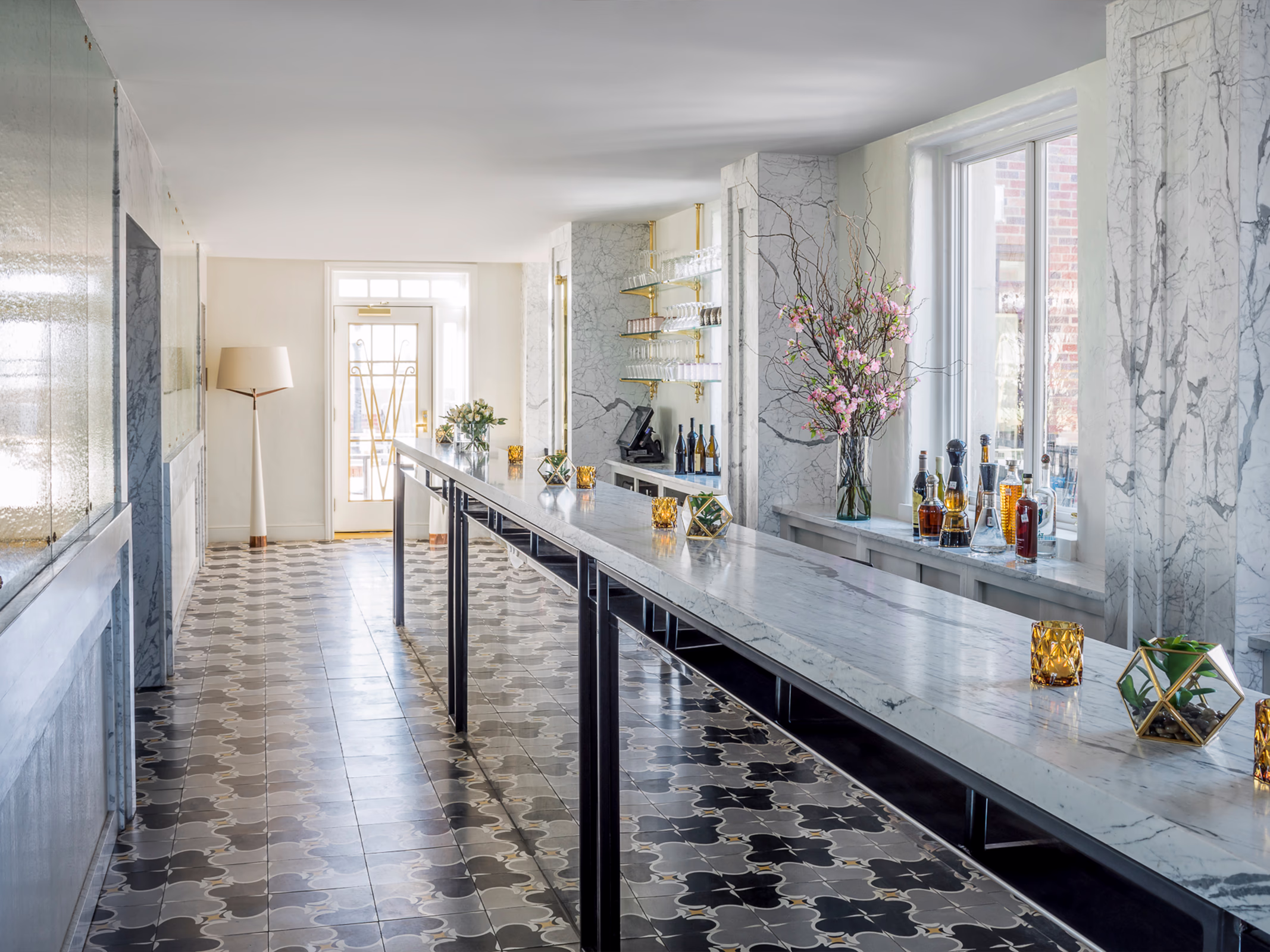 Marble bar at La Perla in El Paso with black metal accents, patterned cement tile flooring, brass glassware shelving, and cherry blossom branches.