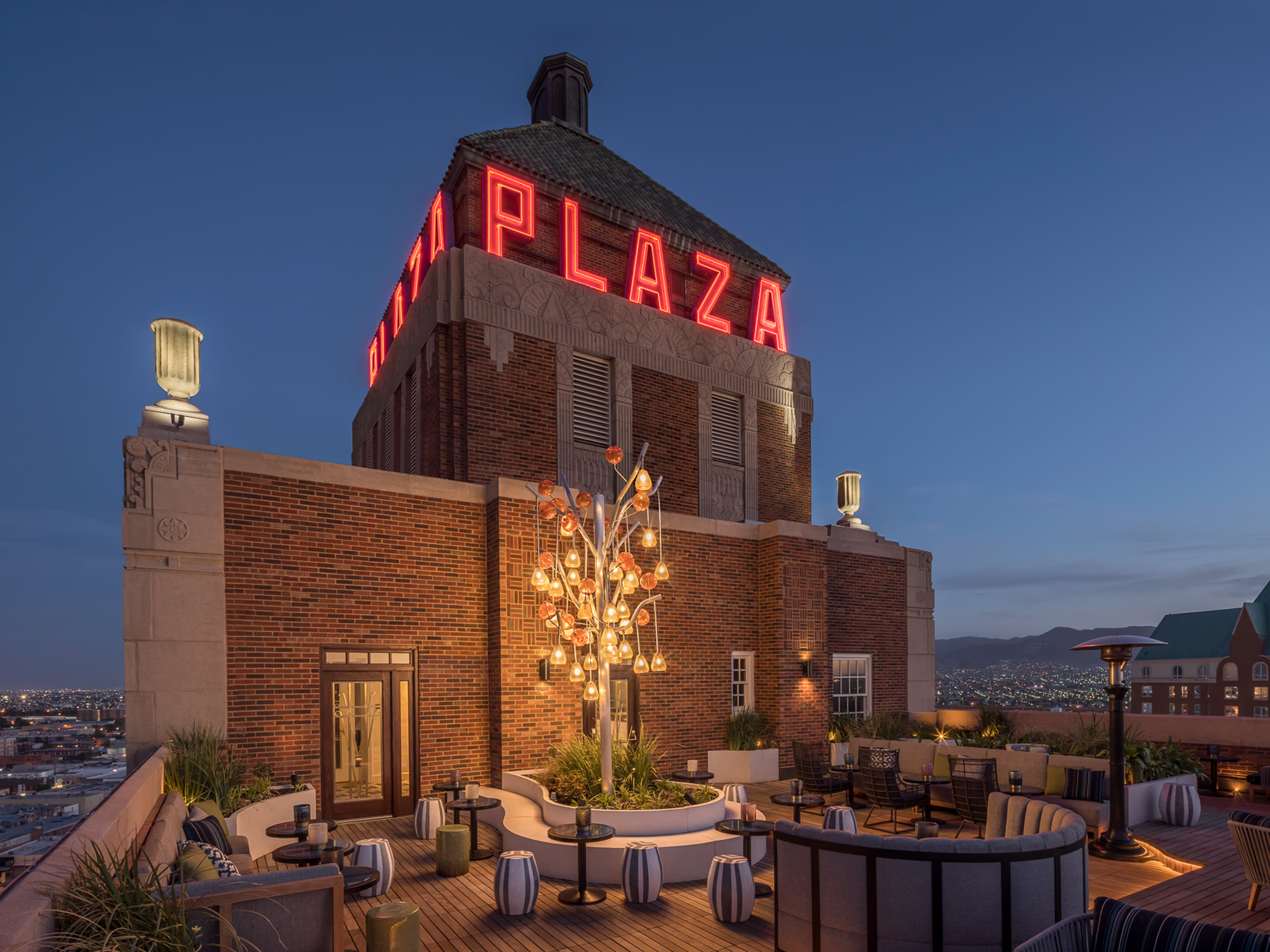 Exterior dusk view of La Perla's rooftop terrace in El Paso beneath the historic Art Deco Plaza tower with its illuminated neon sign and city skyline.