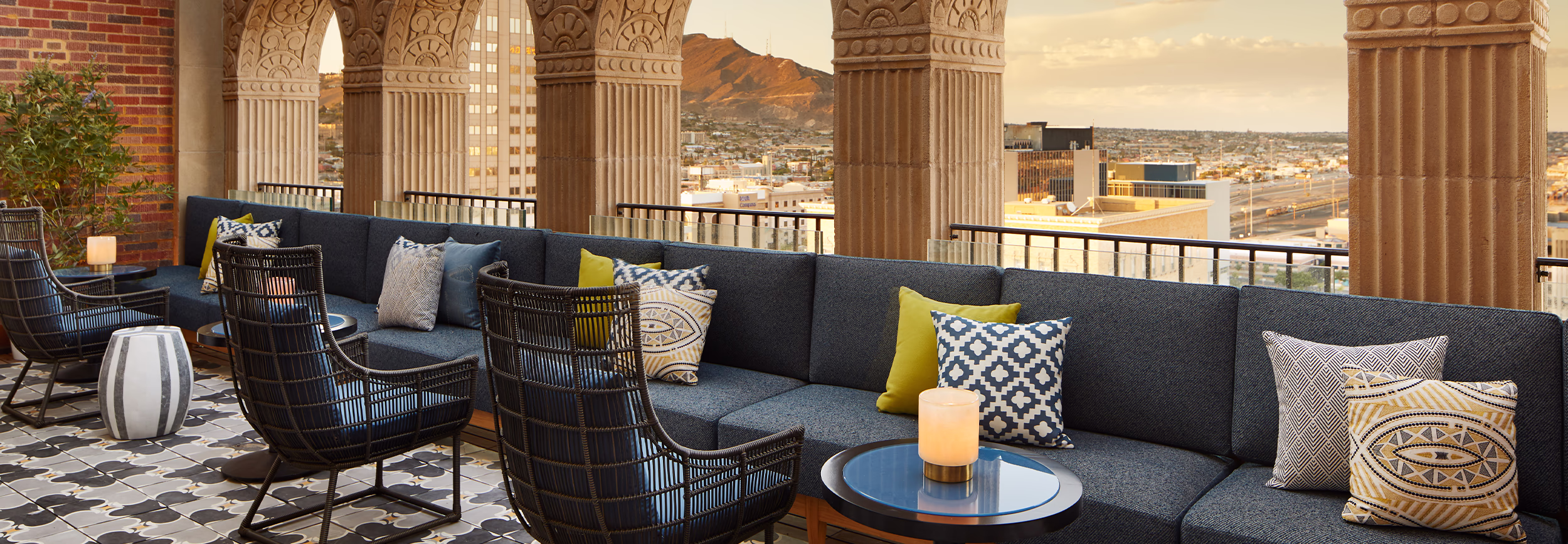 Rooftop terrace at La Perla in El Paso with navy sofas, patterned pillows, and wicker chairs beneath carved arched columns overlooking the mountains at sunset.