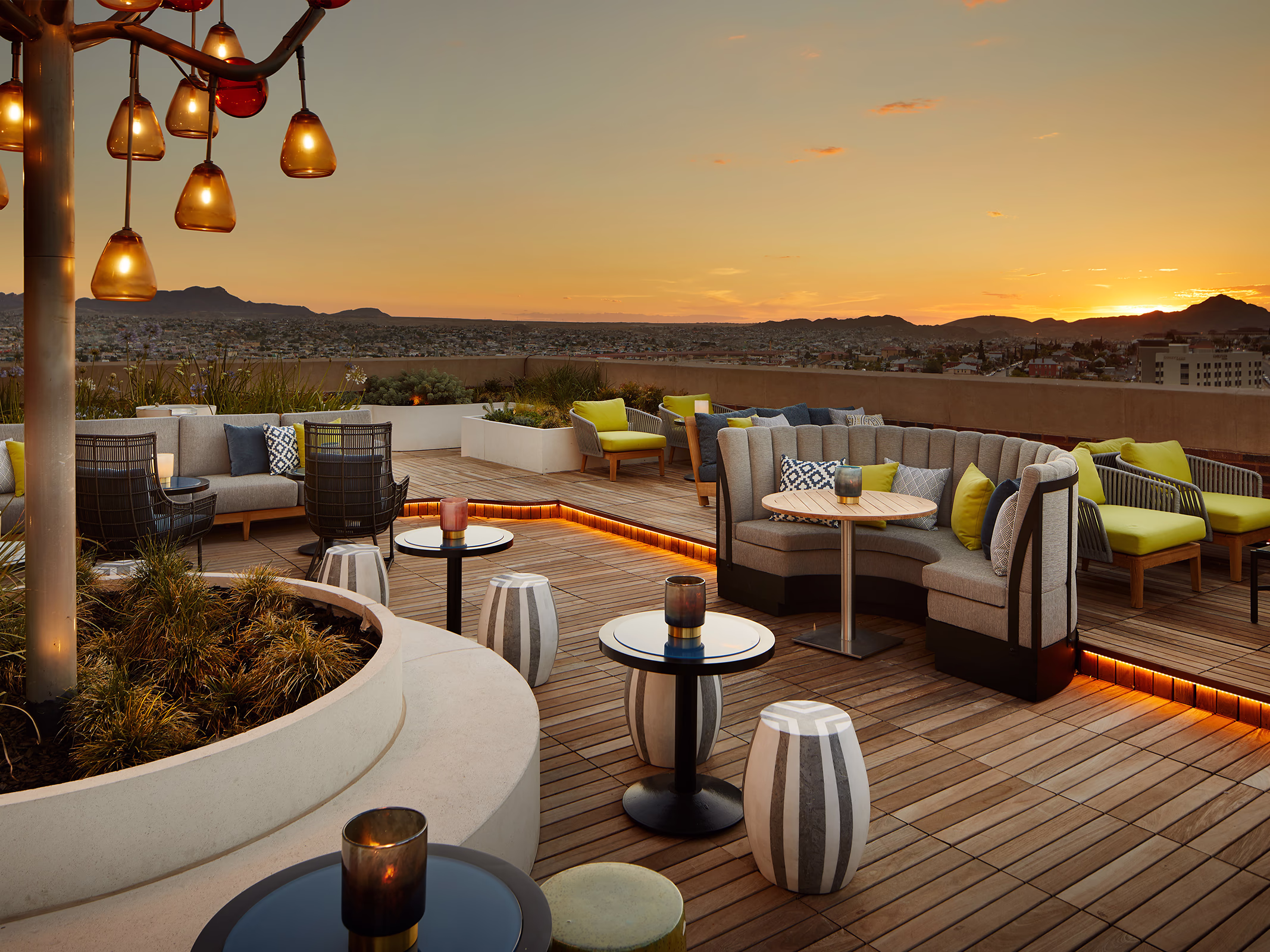 Rooftop terrace at La Perla in El Paso at sunset, with curved gray banquettes, yellow accent chairs, striped side tables, and a custom amber tree-light fixture.