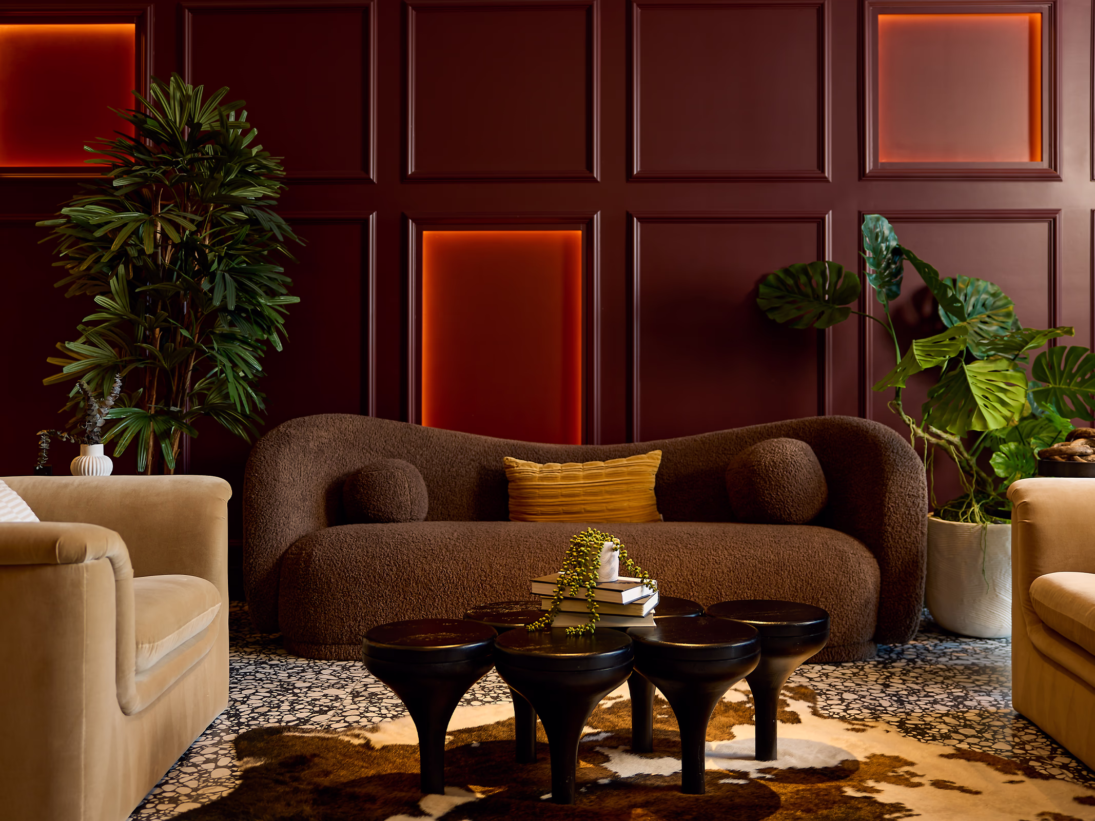 A moody clubhouse lounge vignette at Modera SoBro featuring a curved chocolate boucle sofa, sculptural black coffee tables, and a deep burgundy paneled wall with warm amber backlit niches.