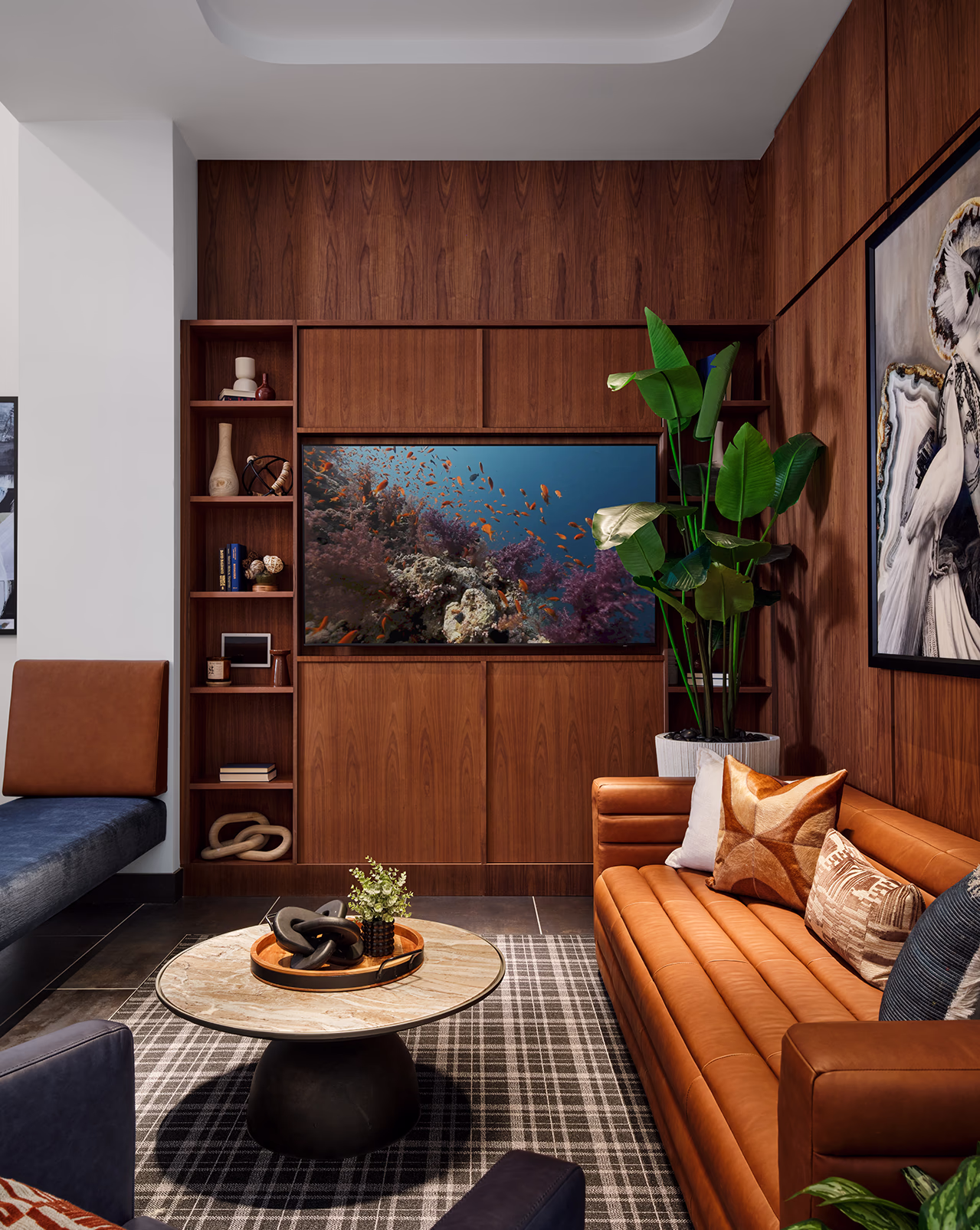 A sophisticated clubhouse lounge at Modera SoBro featuring a cognac leather sofa, walnut built-in shelving, a large TV displaying a coral reef screensaver, and eclectic art and decor.