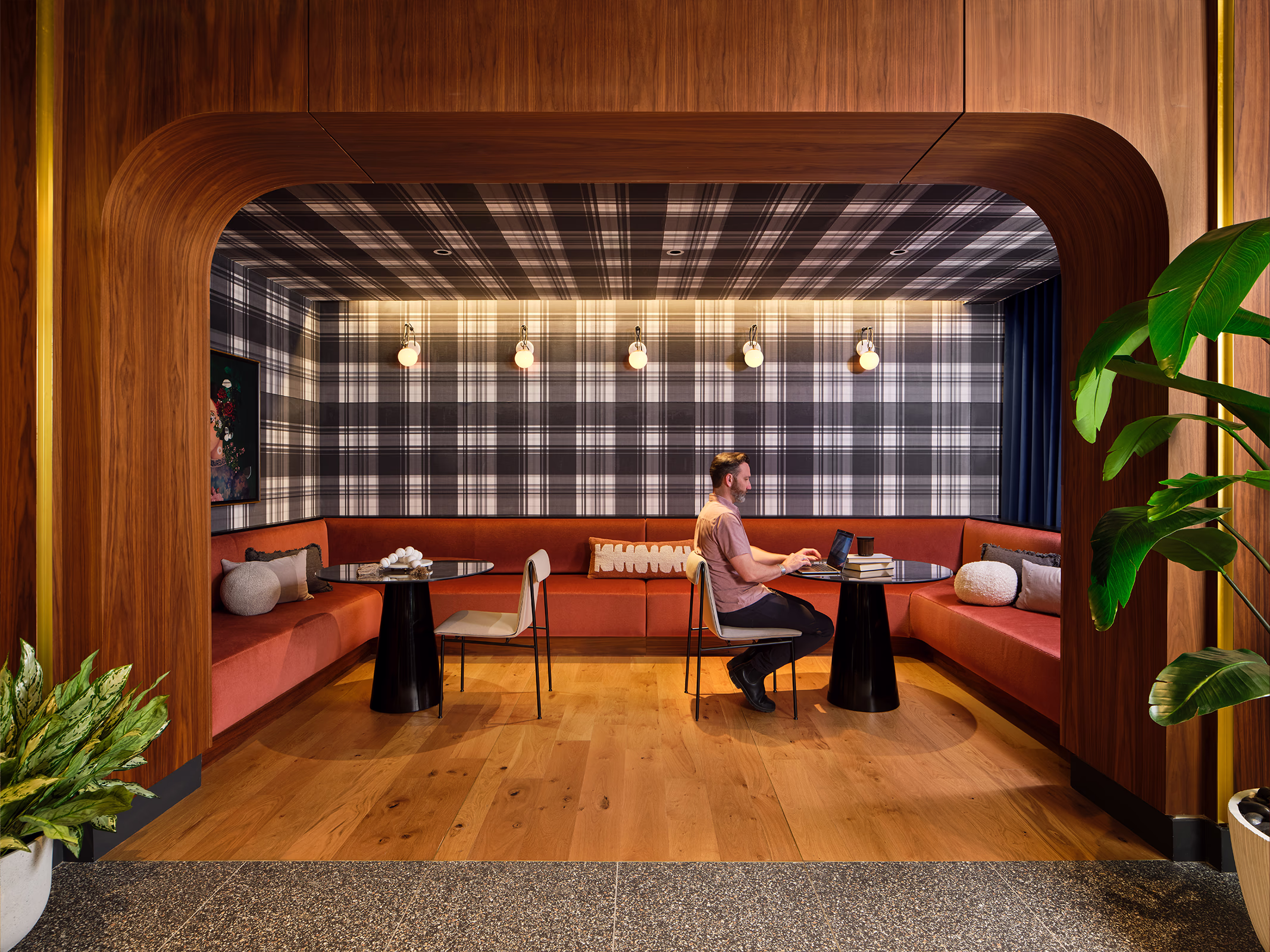 A resident works on a laptop inside a cozy co-working booth at Modera SoBro, framed by a curved walnut archway, plaid wallpaper, and a wraparound rust-red velvet banquette.