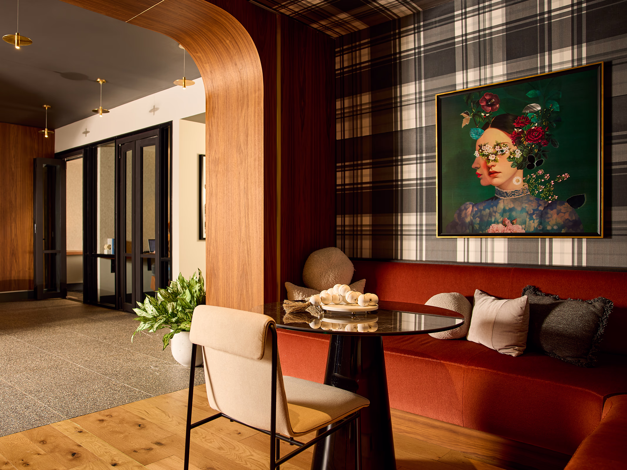 A stylish apartment lobby lounge featuring a rust-red velvet banquette, round black bistro table, plaid wallpaper, and a surrealist floral portrait painting, framed by a curved wood archway.