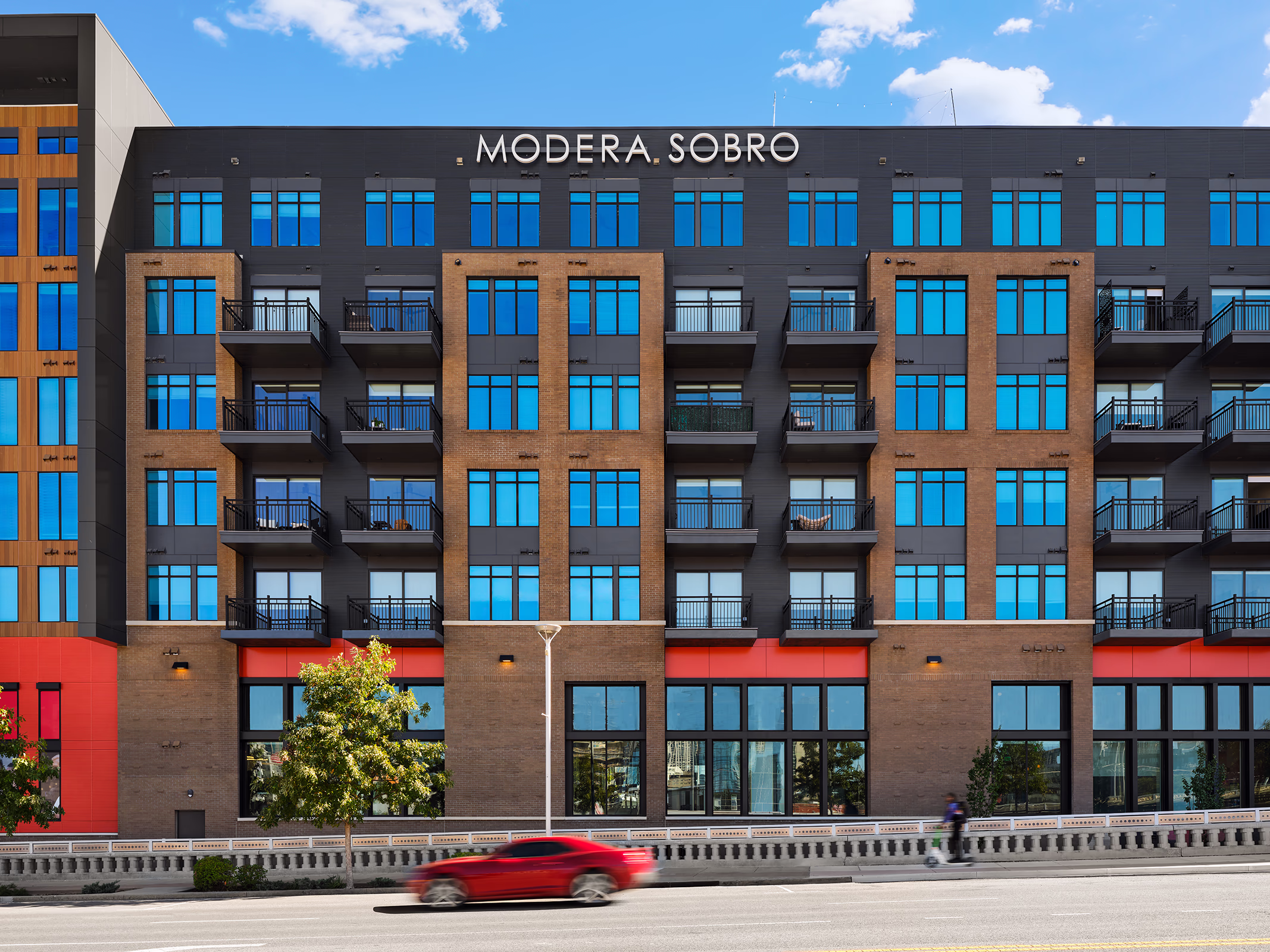 Exterior facade of Modera SoBro, a modern mid-rise apartment building featuring brick cladding, dark gray panels, blue-tinted windows, black balconies, and red retail accents at street level, photographed on a sunny day.