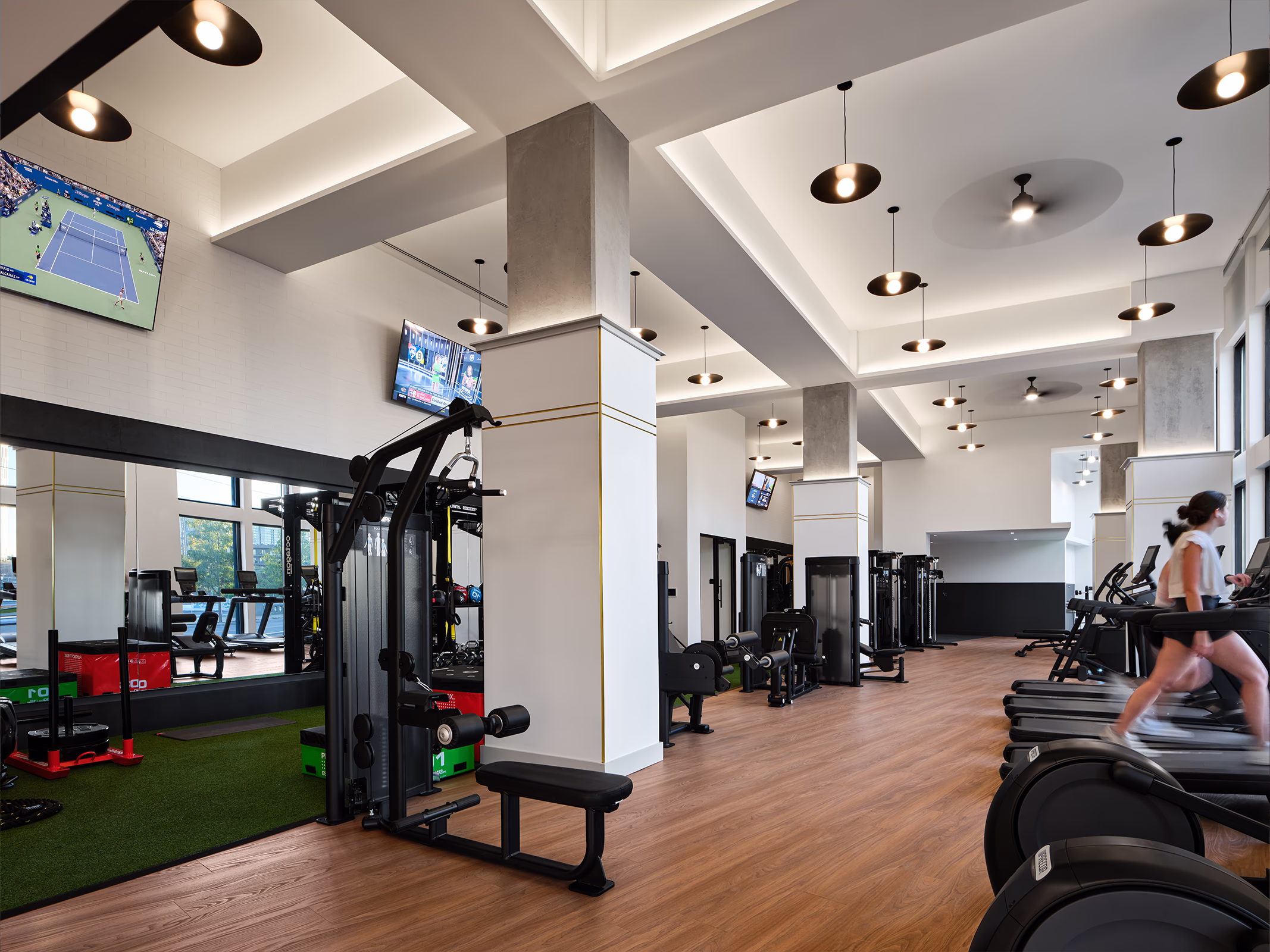 A spacious, well-equipped fitness center at Modera SoBro featuring strength and cardio machines, an artificial turf training zone, multiple TVs, and stylish pendant lighting in a bright open space.