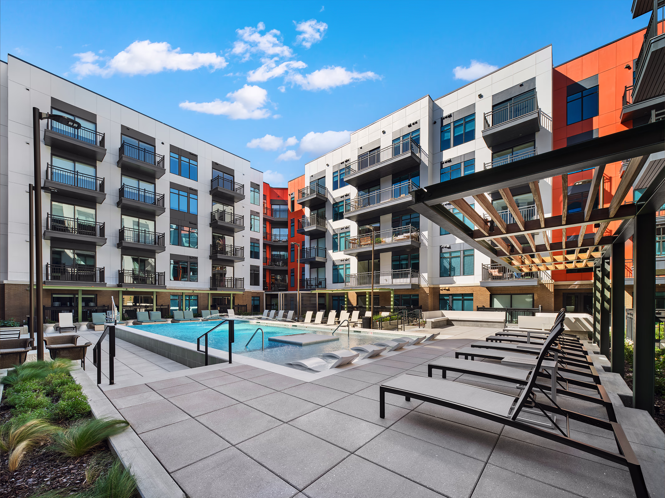 Modera SoBro's resort-style outdoor pool courtyard surrounded by multi-story residential buildings with red and white facades, black balconies, lounge chairs, and a pergola structure under a blue sky.