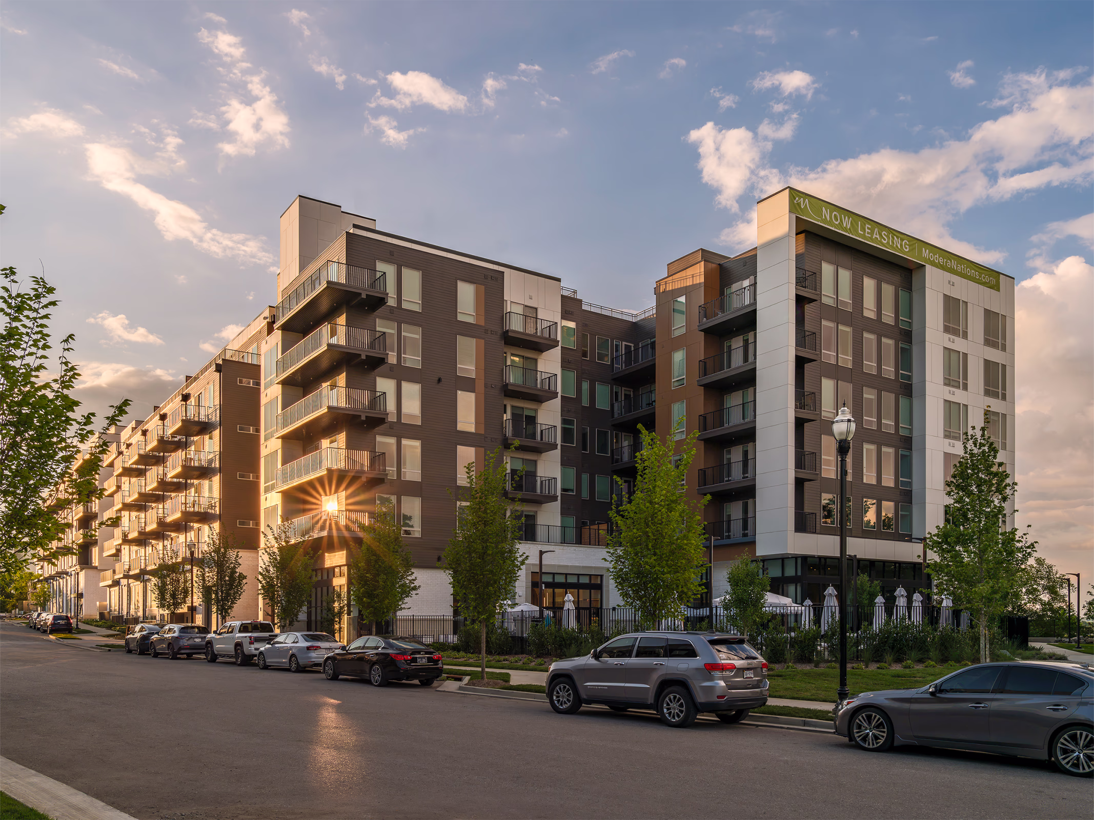 Modera Nations apartments in Nashville at golden hour, with sun glowing through stacked balconies above a tree-lined street and a fenced umbrella patio.