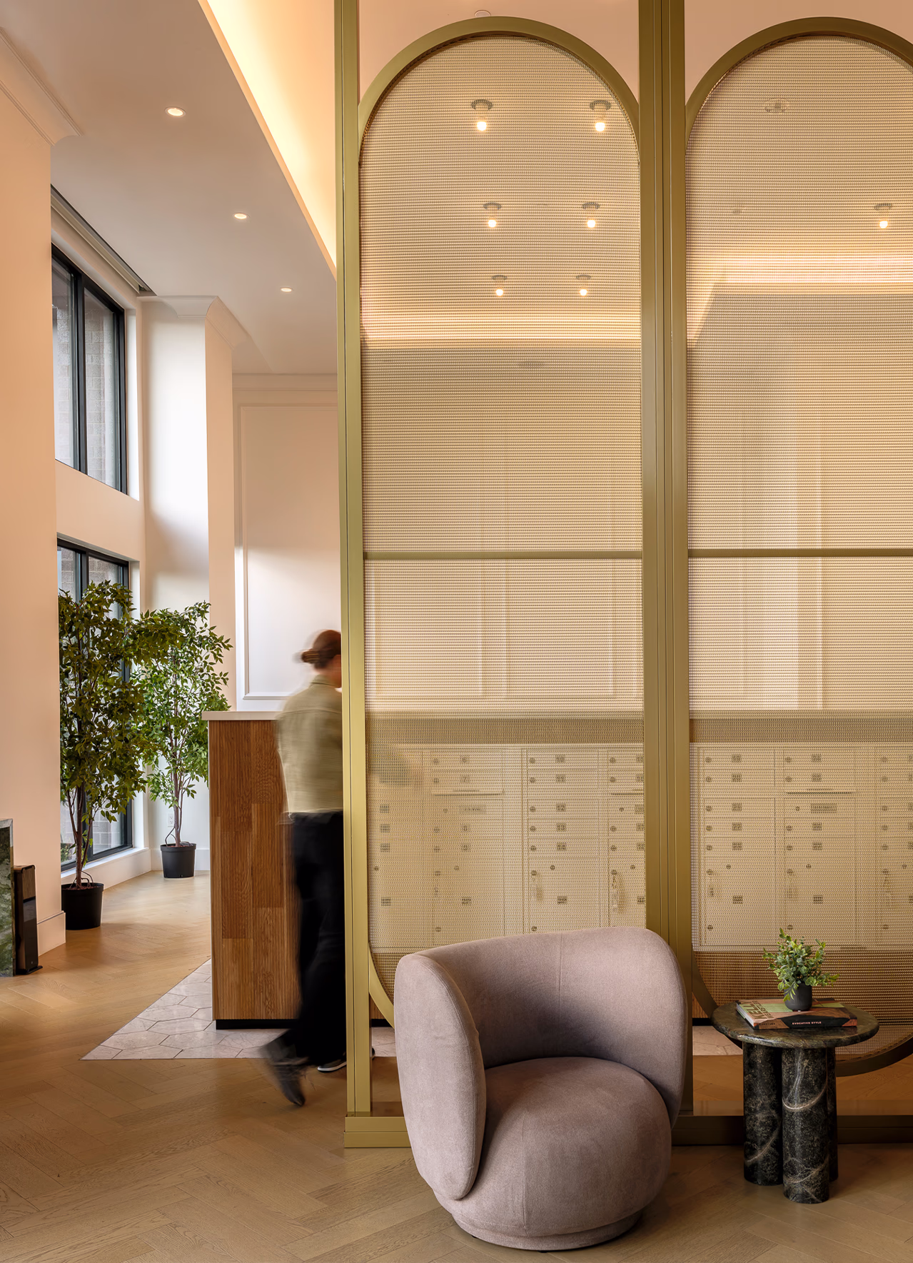 Modera Nations mailroom in Nashville behind arched brass-framed mesh screens, with a curved lounge chair, marble side table and herringbone wood floors.