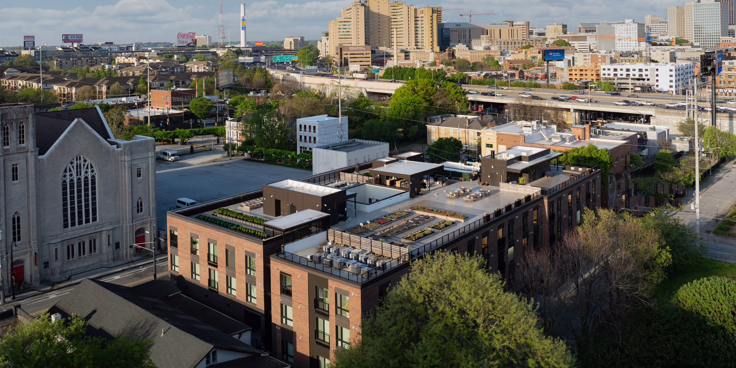 Aerial daytime view of the Front Porch development showing the rooftop community gardens and event spaces atop the Phase I co-living building, with a historic church to the left and the Atlanta skyline in the background.