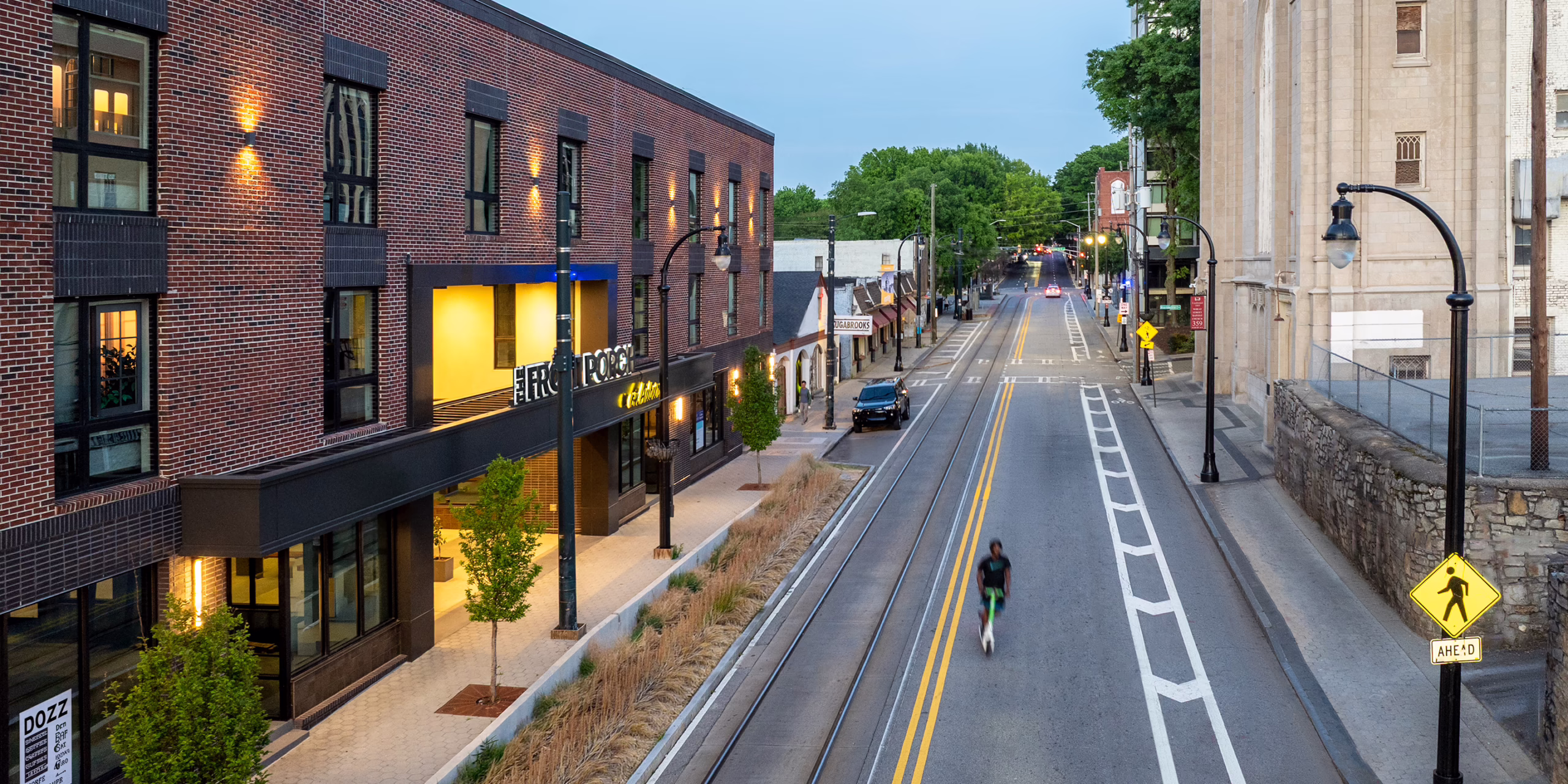 Street-level dusk view of the Front Porch development along Auburn Avenue, showing the illuminated retail entrance and signage, streetcar tracks, landscaped sidewalk, and a cyclist passing by, with the Haugabrooks building visible in the distance.