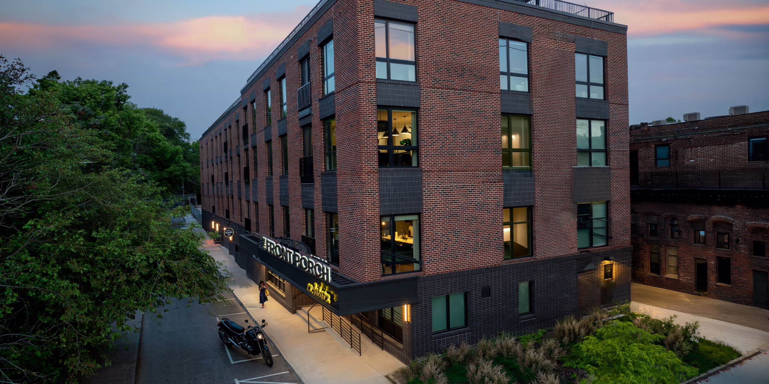 Elevated dusk view of the Front Porch building exterior showing the illuminated entrance canopy with signage, warm light glowing through residential windows, and brick facade of the co-living building in the Martin Luther King Jr. National Historic District.