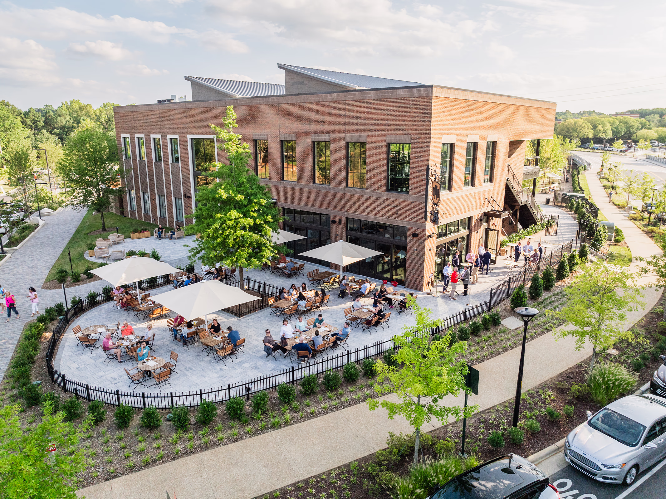 Aerial view of a two-story brick restaurant at The Bowl at Ballantyne in Charlotte, with a busy fenced dining patio, umbrellas and landscaped walkways.