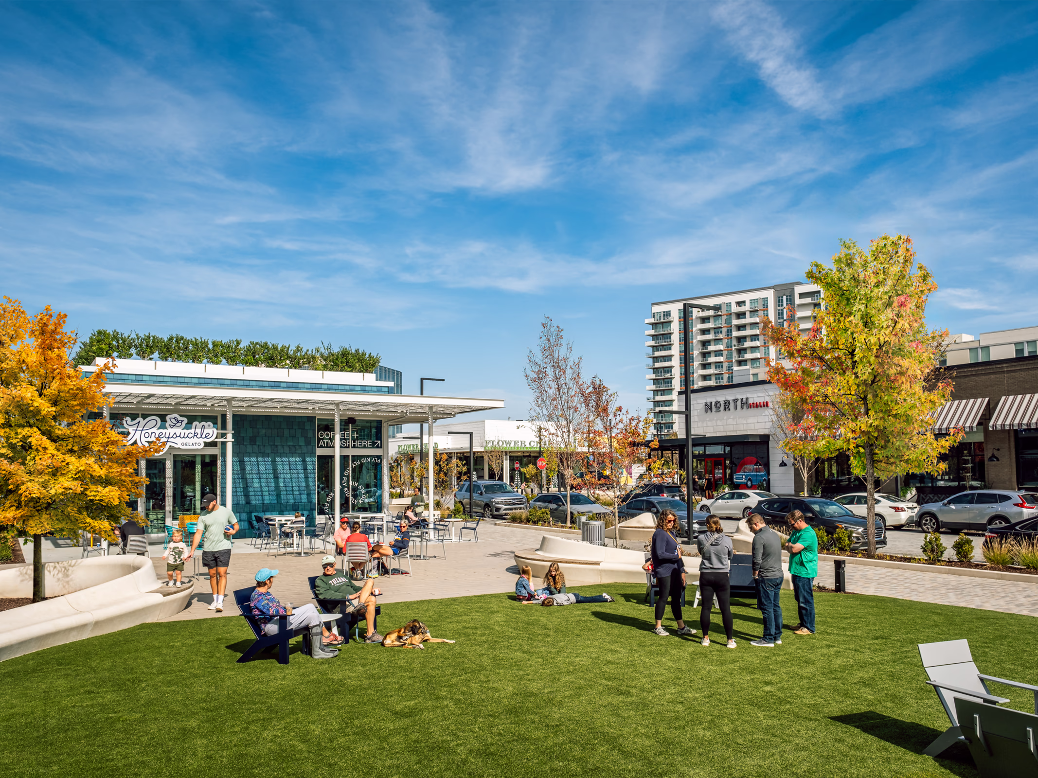 Central lawn at The Bowl at Ballantyne in Charlotte, with people relaxing on the grass near Honeysuckle Gelato, curved benches and main street retail.