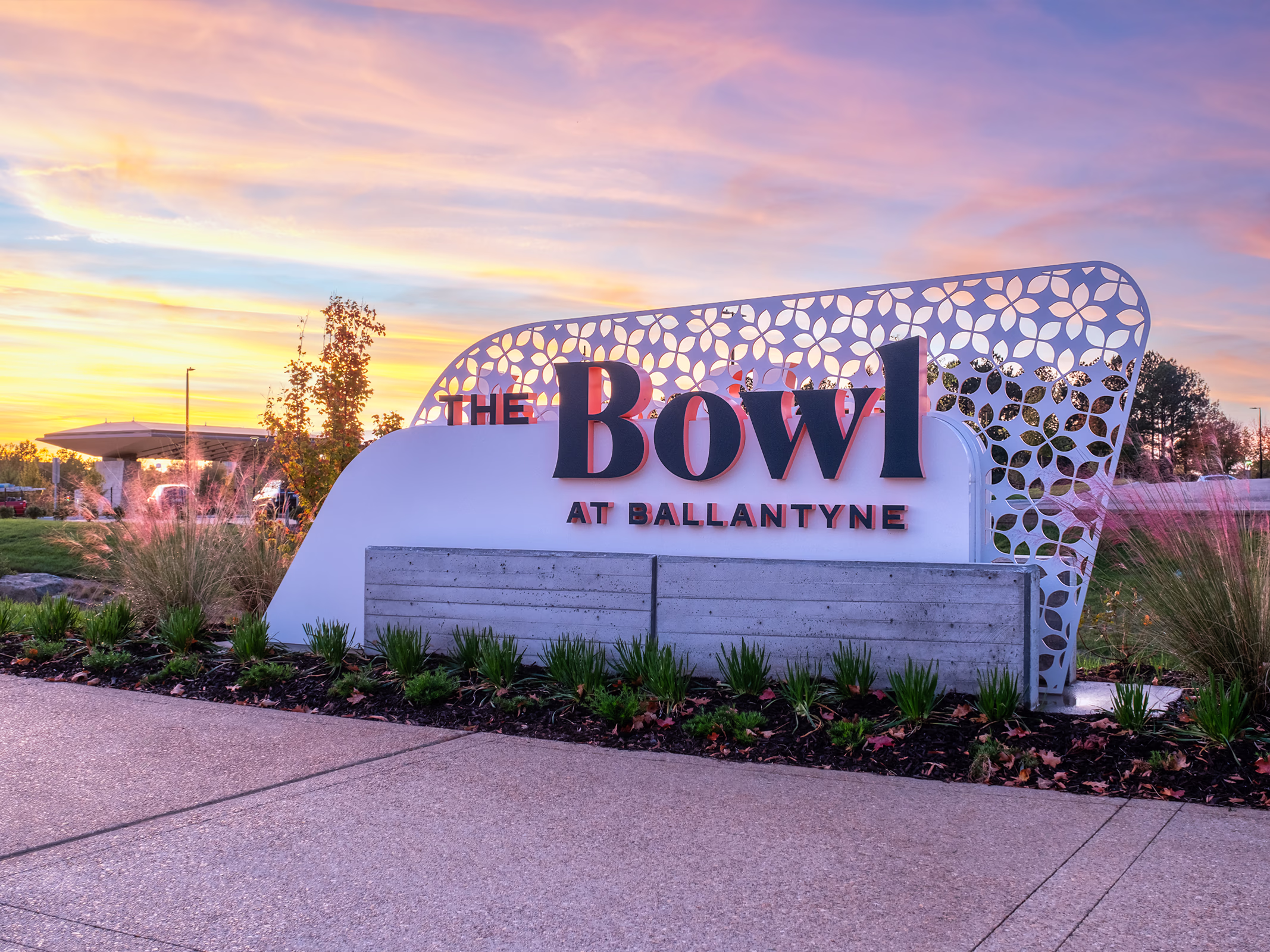 The Bowl at Ballantyne entry monument sign in Charlotte at sunset, with a perforated white metal panel, board-formed concrete base and native grasses.