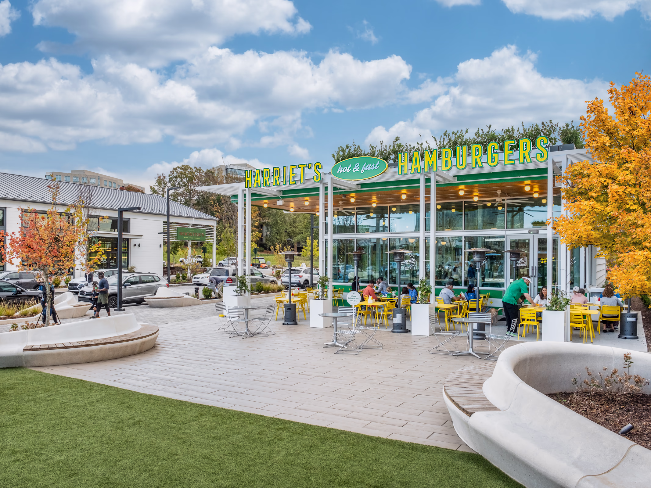 Harriet's Hamburgers at The Bowl at Ballantyne in Charlotte, with a pergola-covered patio, yellow cafe seating, curved plaza benches and fall trees.