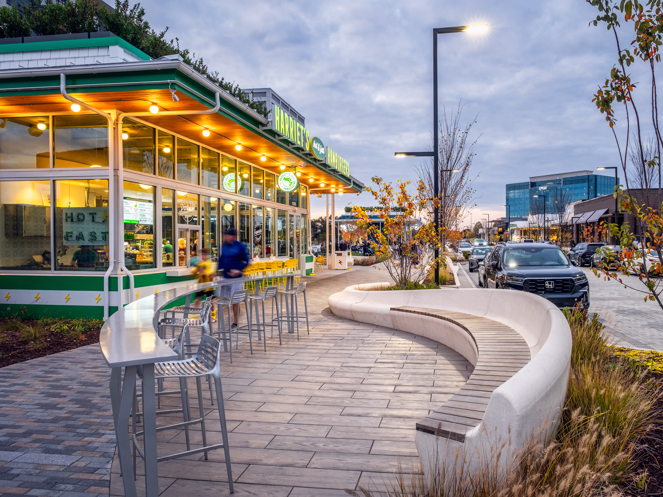 Harriet's Hamburgers at The Bowl at Ballantyne in Charlotte at dusk, with outdoor bar seating, a curved wood bench and a landscaped main street walk.