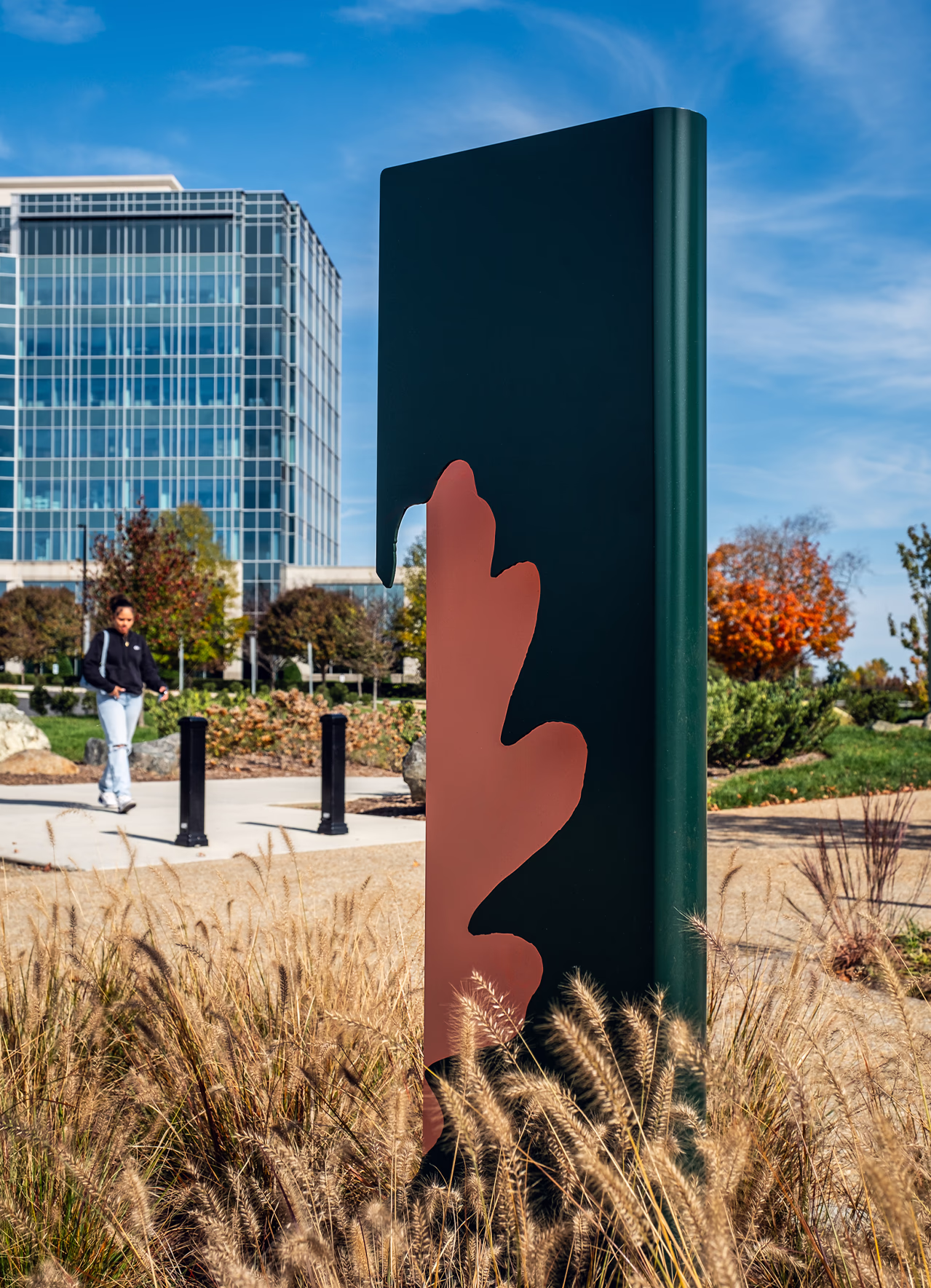 Dark green wayfinding sign with an oak leaf cutout at The Bowl at Ballantyne in Charlotte, set among ornamental grasses near a glass office tower.