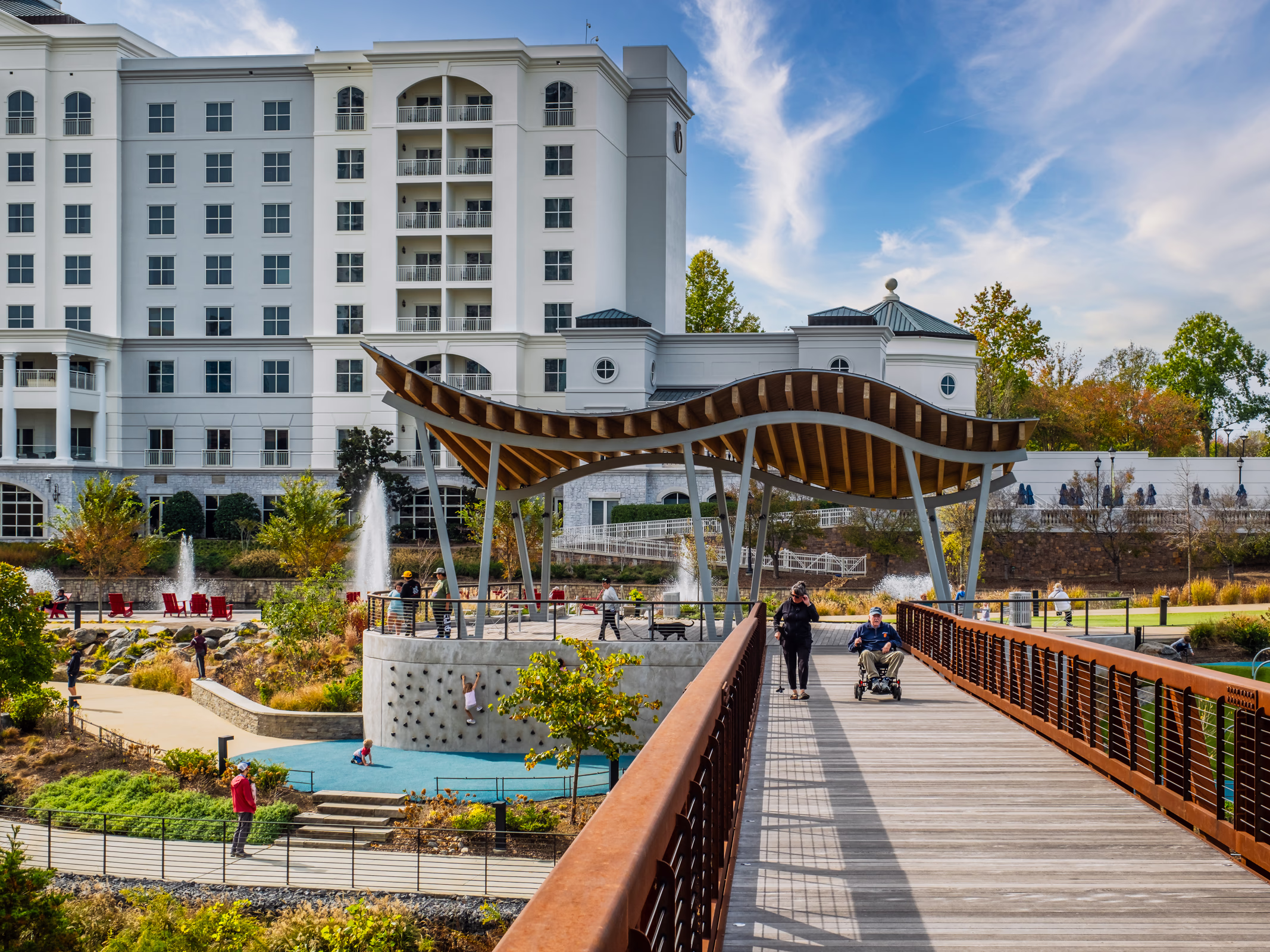 Pedestrian bridge at The Bowl at Ballantyne in Charlotte leading to a wave-roofed timber pavilion, a kids' climbing wall, fountains and landscaped park.