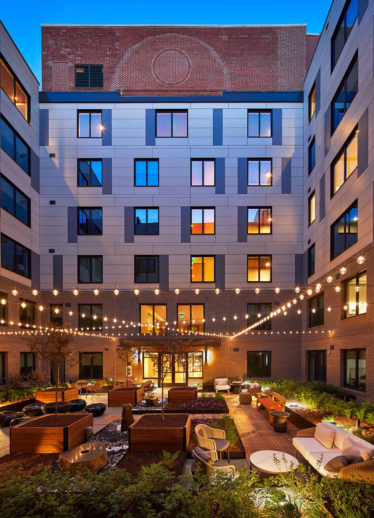 Evening view of CityHouse Old Town's six-story open-air atrium courtyard in Alexandria, Virginia, illuminated by string lights, with landscaped planters, lounge seating, wood decking, and residential windows glowing above.