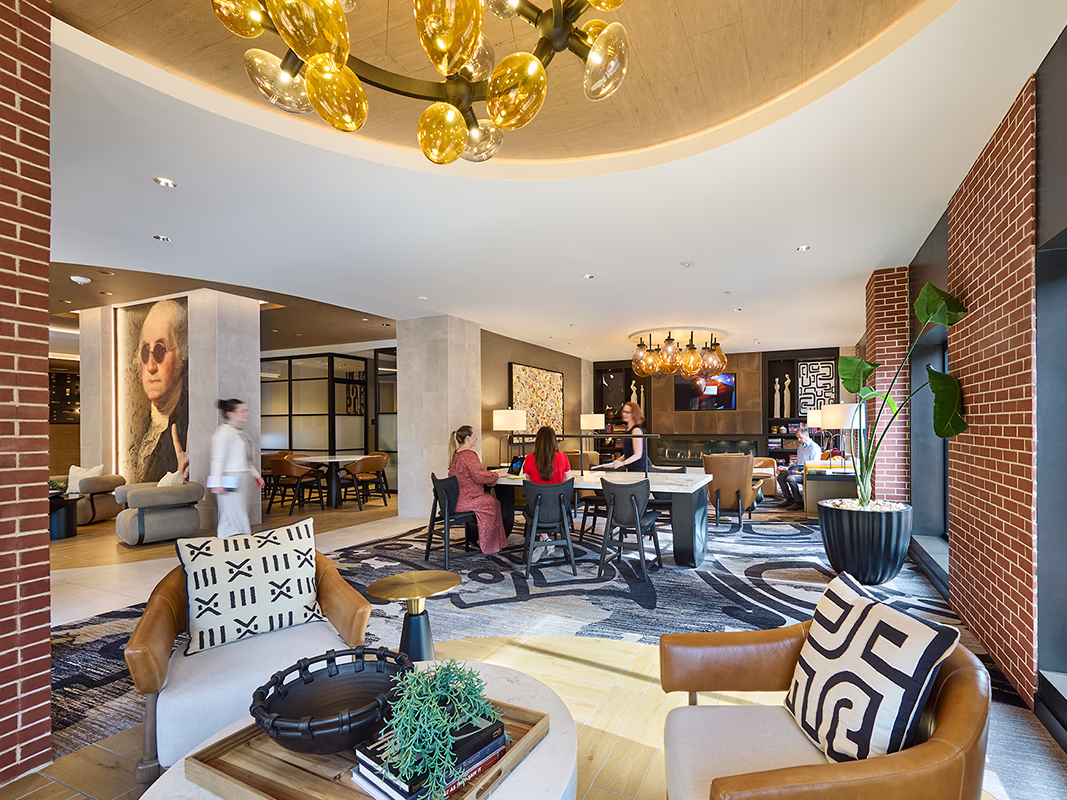 Private club lounge at CityHouse Old Town in Alexandria, Virginia, with globe chandelier lighting, a gas fireplace, communal work table, leather lounge seating, patterned rug, and colonial portrait art visible in the background.