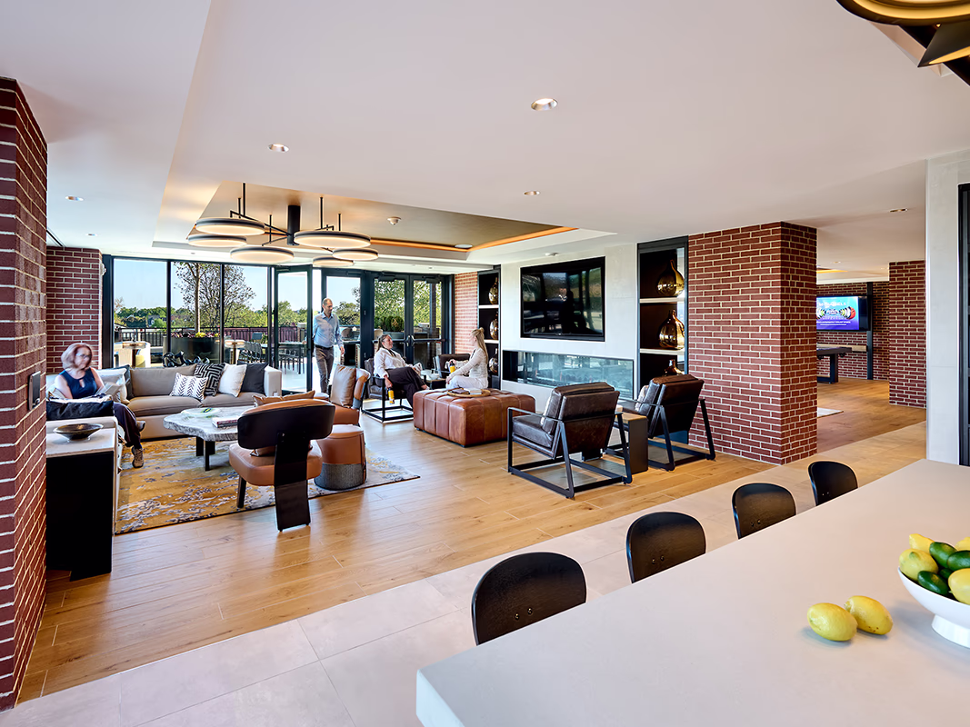 Private club suite at CityHouse Old Town in Alexandria, Virginia, with a fireplace, lounge and leather club chair seating, TV, exposed brick columns, kitchen island, and doors opening to a rooftop terrace with sunset views.
