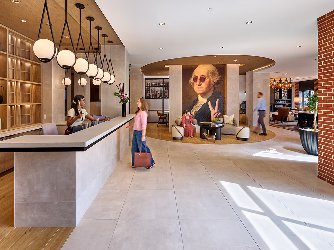 Concierge desk and lobby lounge at CityHouse Old Town in Alexandria, Virginia, with globe pendant lighting, a resident checking in, seating around a playful George Washington portrait, and views into the coworking area beyond.