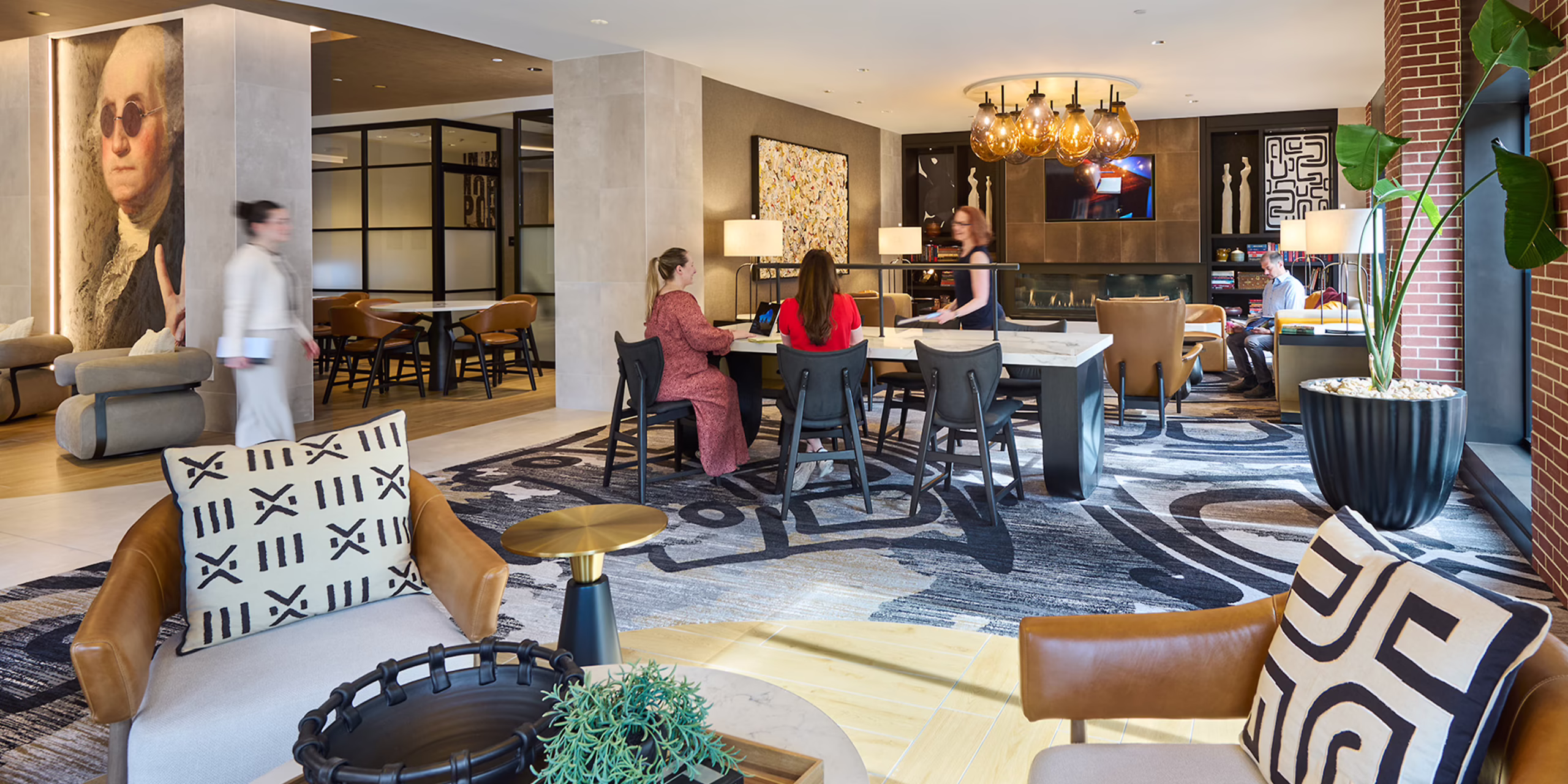 Coworking and lounge space at CityHouse Old Town in Alexandria, Virginia, with a communal work table, gas fireplace, patterned area rug, leather lounge chairs, and oversized colonial portrait art beneath globe pendant lighting.
