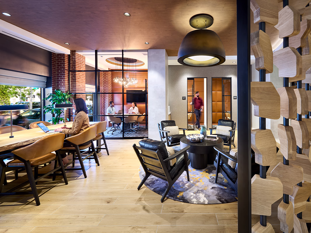 Coworking space at CityHouse Old Town in Alexandria, Virginia, with communal work tables, leather lounge seating around a round coffee table, decorative wood-carved room divider, and glass-walled private meeting rooms in the background.