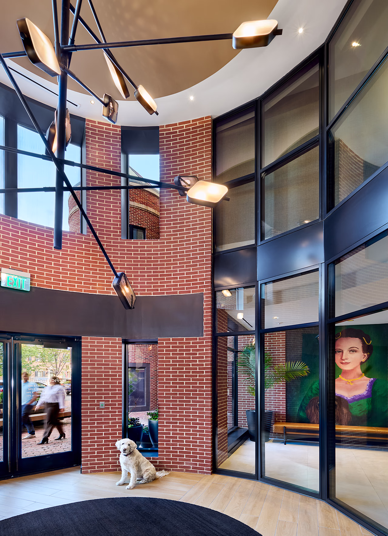 Double-height glass and brick entrance vestibule at CityHouse Old Town in Alexandria, Virginia, featuring a sculptural chandelier, exposed brick walls, colorful portrait art, and a friendly dog seated on the wood floor.