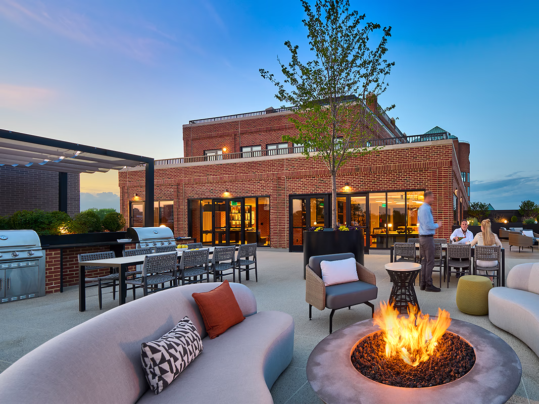 Evening view of CityHouse Old Town's rooftop social terrace in Alexandria, Virginia, with a gas fire pit, curved lounge sofa, grilling stations, communal dining tables, and residents socializing at dusk.