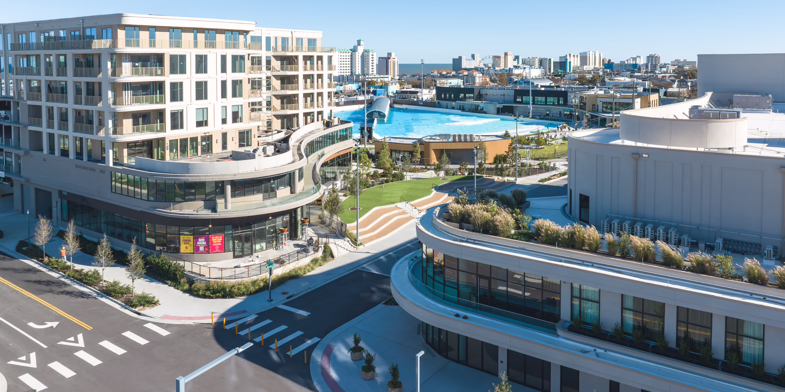 Aerial view of Atlantic Park mixed-use district in Virginia Beach, VA, featuring curved residential and retail buildings, a public plaza, and the WaveGarden surf lagoon with the oceanfront skyline in the background.
