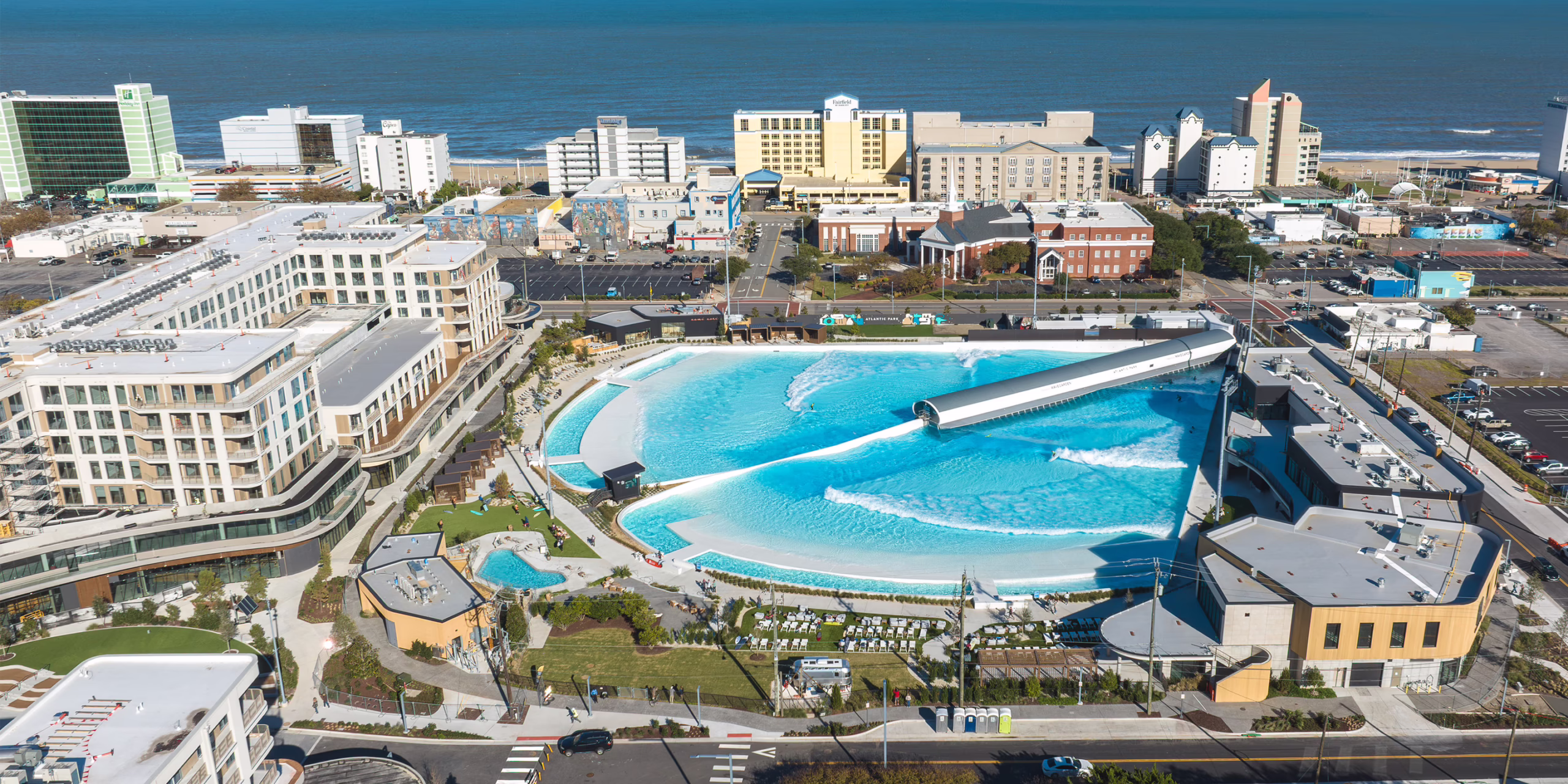 Aerial view of Atlantic Park's WaveGarden surf lagoon in Virginia Beach, VA, showing surfers riding waves alongside urban residential buildings, boardwalk hotels, and the Atlantic Ocean.