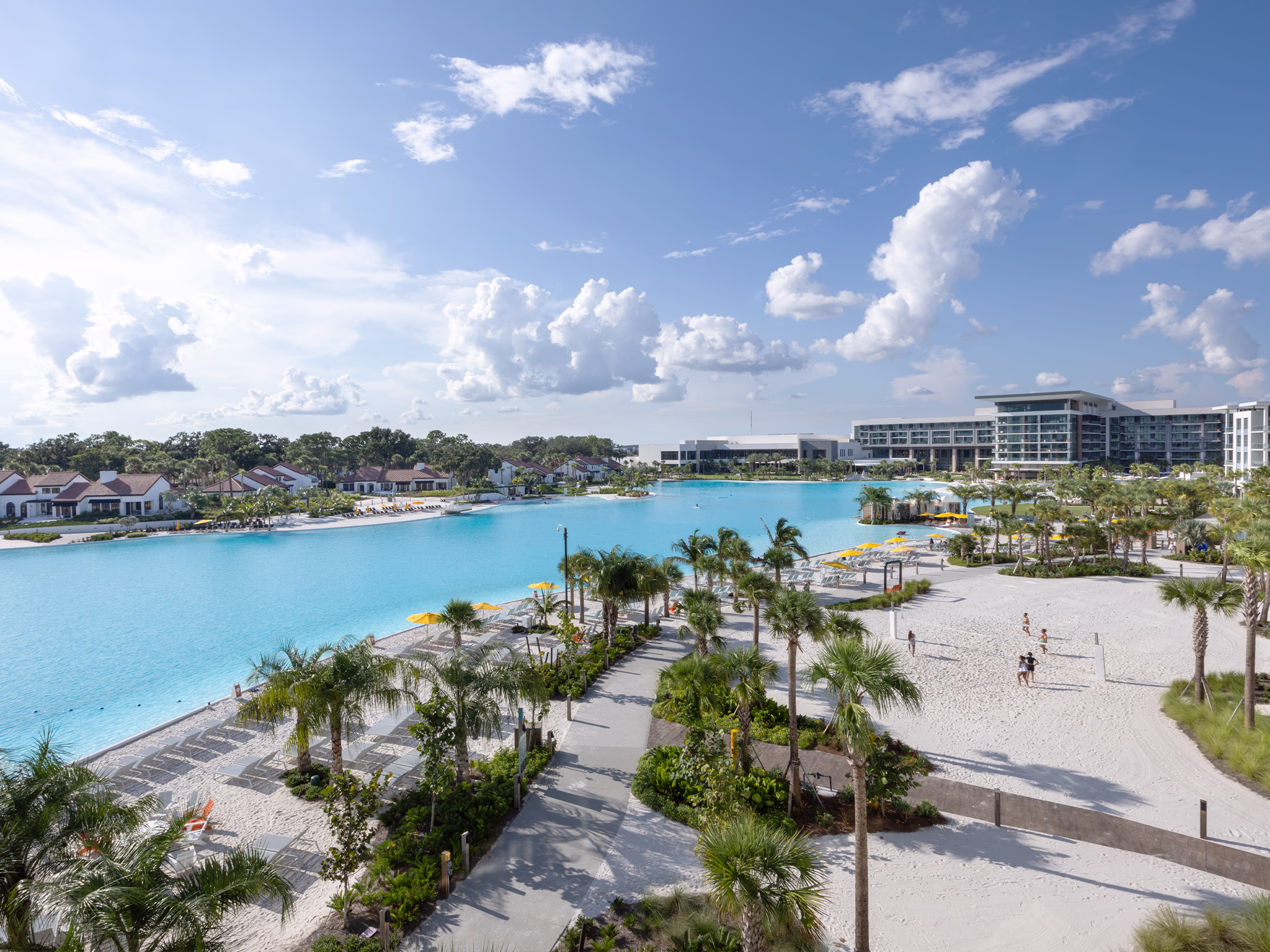 Elevated view of the 8-acre Evermore Bay Crystal Lagoon with white sand beach, palms and yellow umbrellas, Conrad Orlando rising beyond.