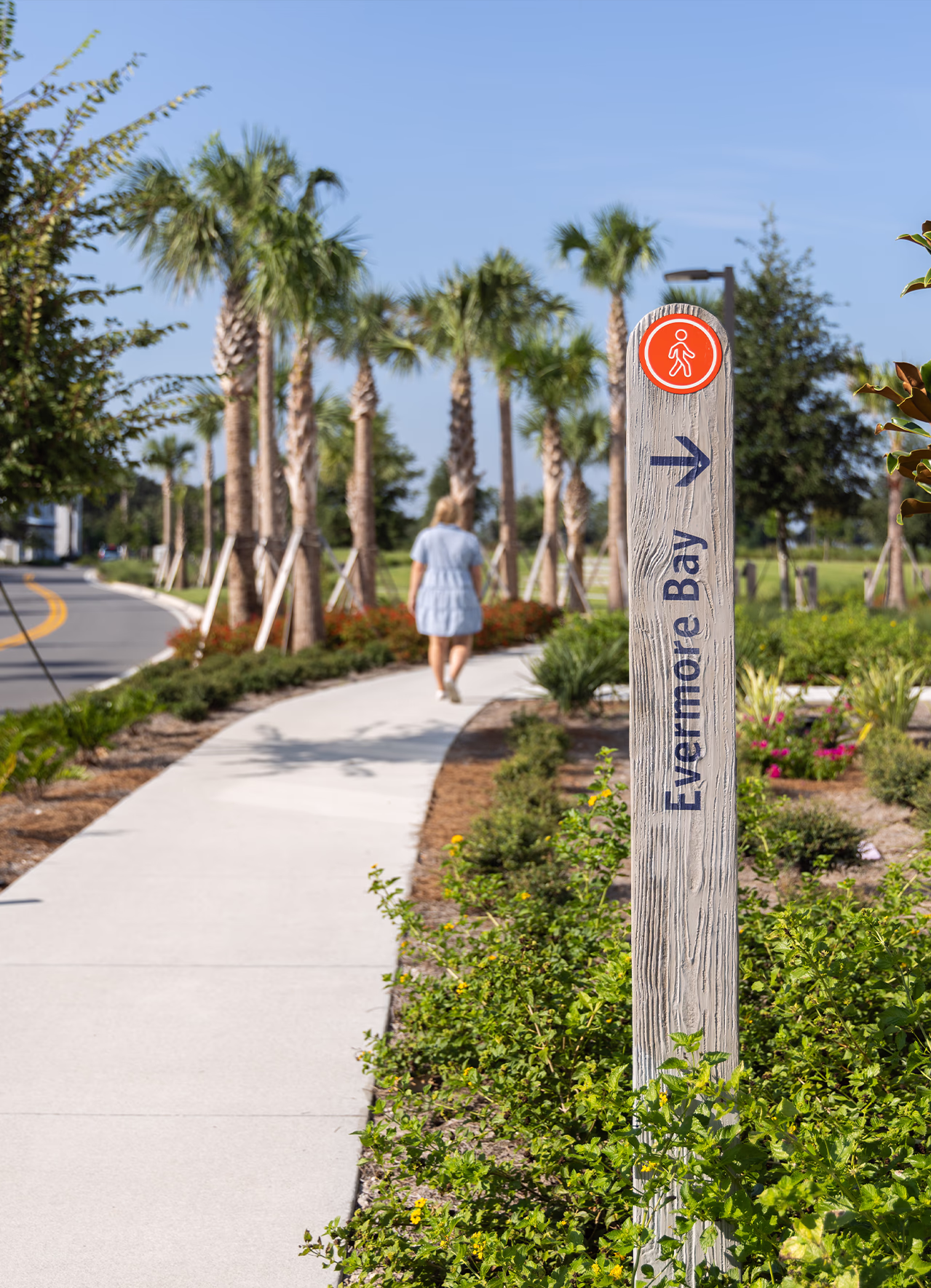 Wood-textured pedestrian marker post with an orange walker icon directing guests to Evermore Bay along a palm-lined resort walking path.