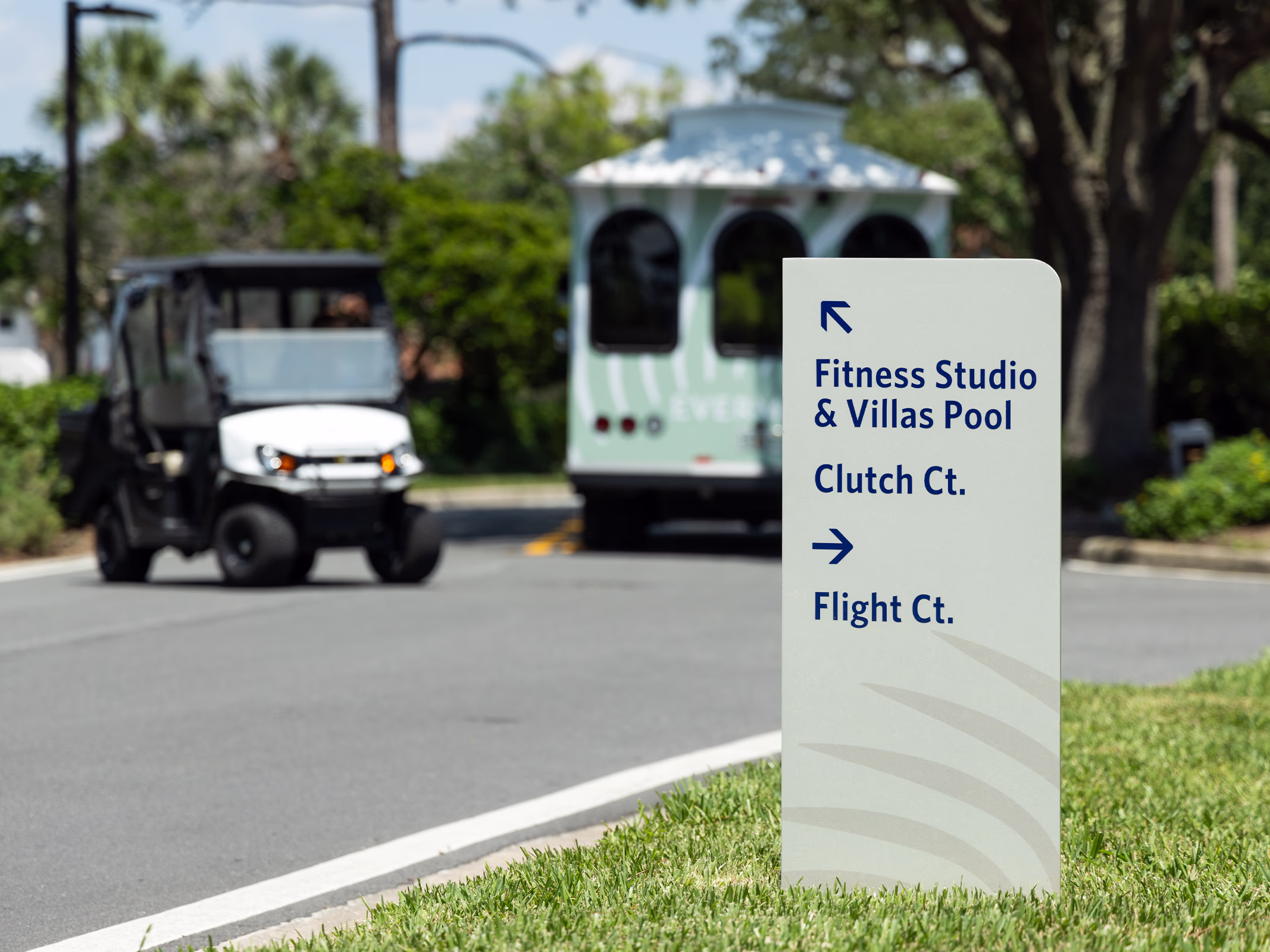 Roadside directional sign with navy arrows to Fitness Studio, Villas Pool, Clutch Ct. and Flight Ct. as a golf cart and resort trolley pass by.