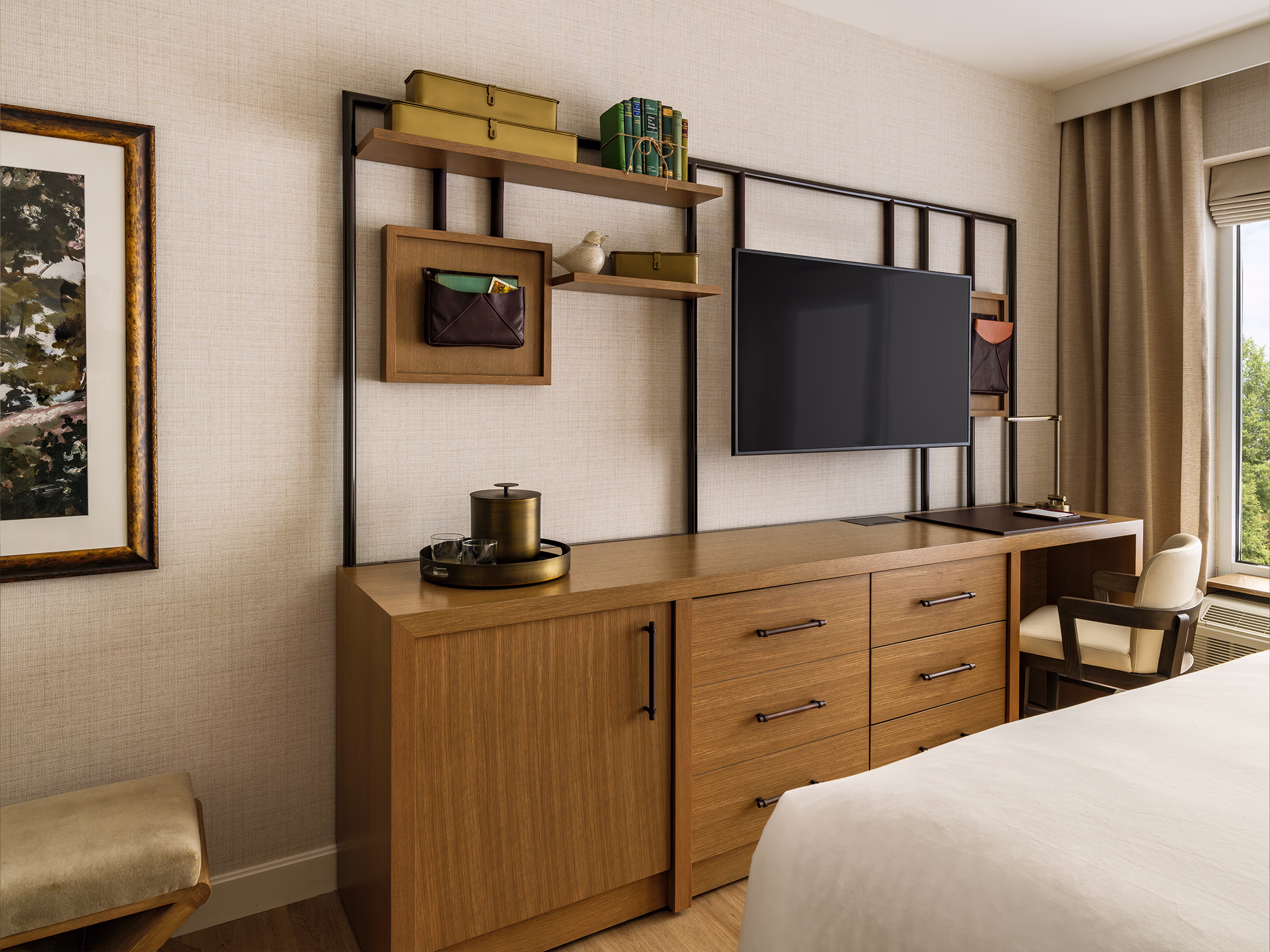 Guestroom media wall at The James F. Martin Inn with wall-mounted TV, wood shelving, brass ice bucket, and curated leather accessories atop a wood dresser.