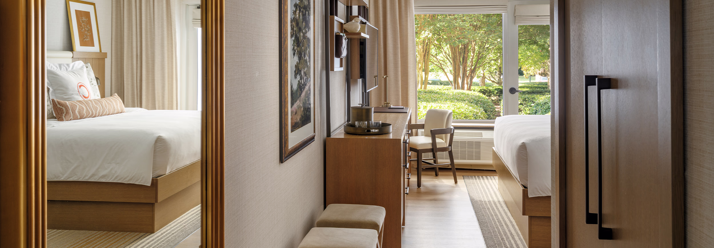 Queen/queen guestroom at The James F. Martin Inn at Clemson University, featuring a gold-framed mirror, wood dresser, and a tree-lined window view.