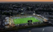 Aerial nighttime view of Centreville Bank Stadium filled with fans, with illuminated signage and lighting, the river in the foreground, and the town visible in the background.
