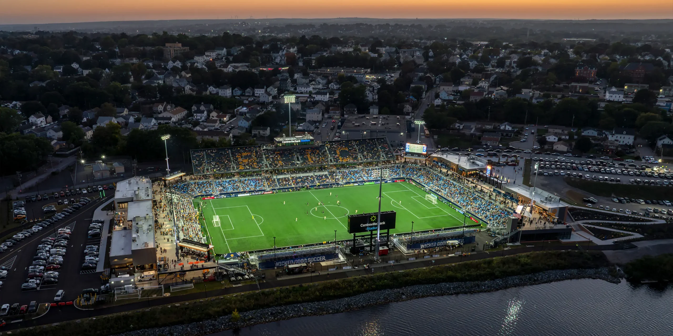 Aerial nighttime view of Centreville Bank Stadium filled with fans, with illuminated signage and lighting, the river in the foreground, and the town visible in the background.
