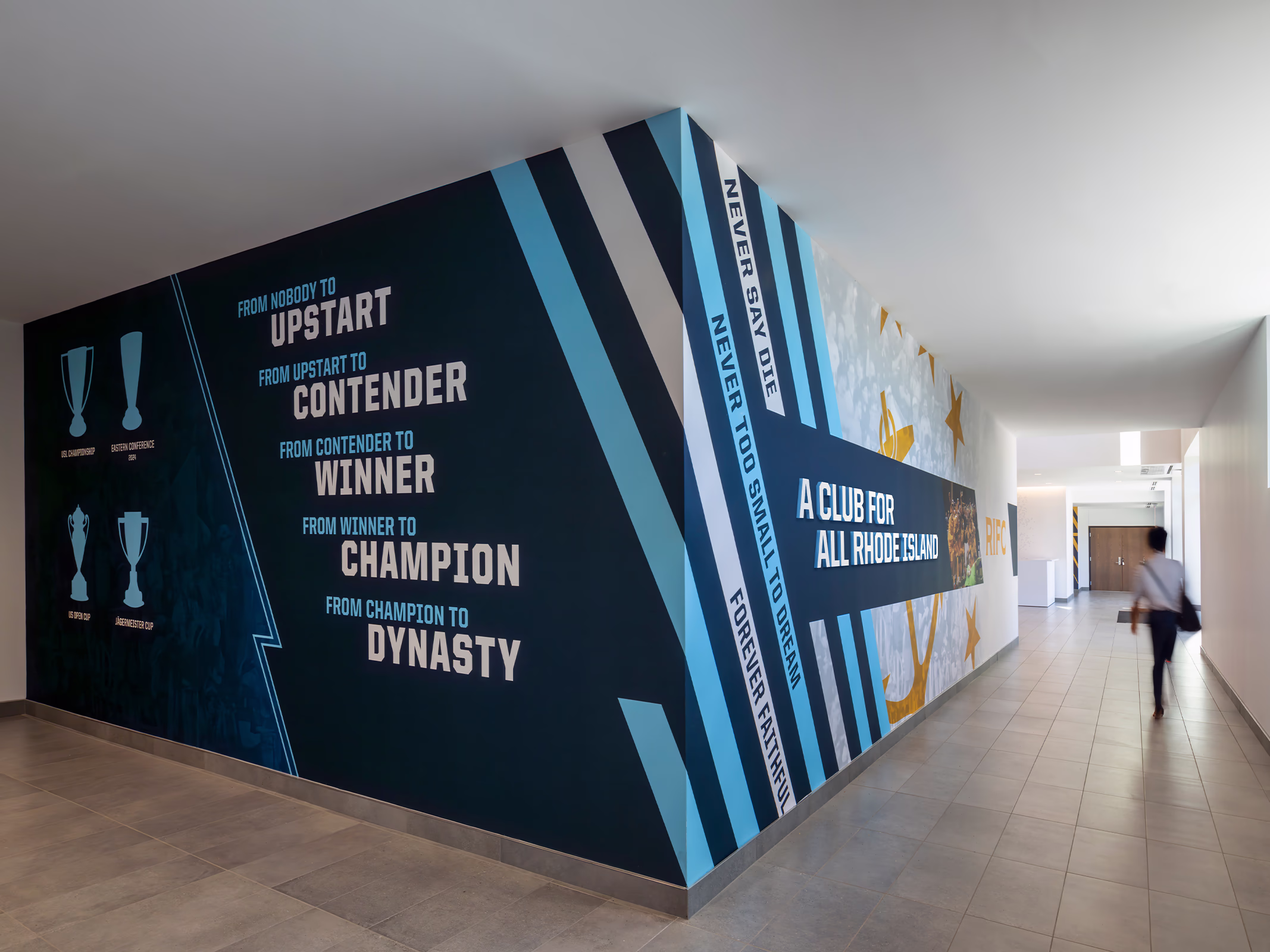 Bold typographic wall graphics with Rhode Island FC mottos and trophy icons wrap a back-of-house corridor at Centreville Bank Stadium.