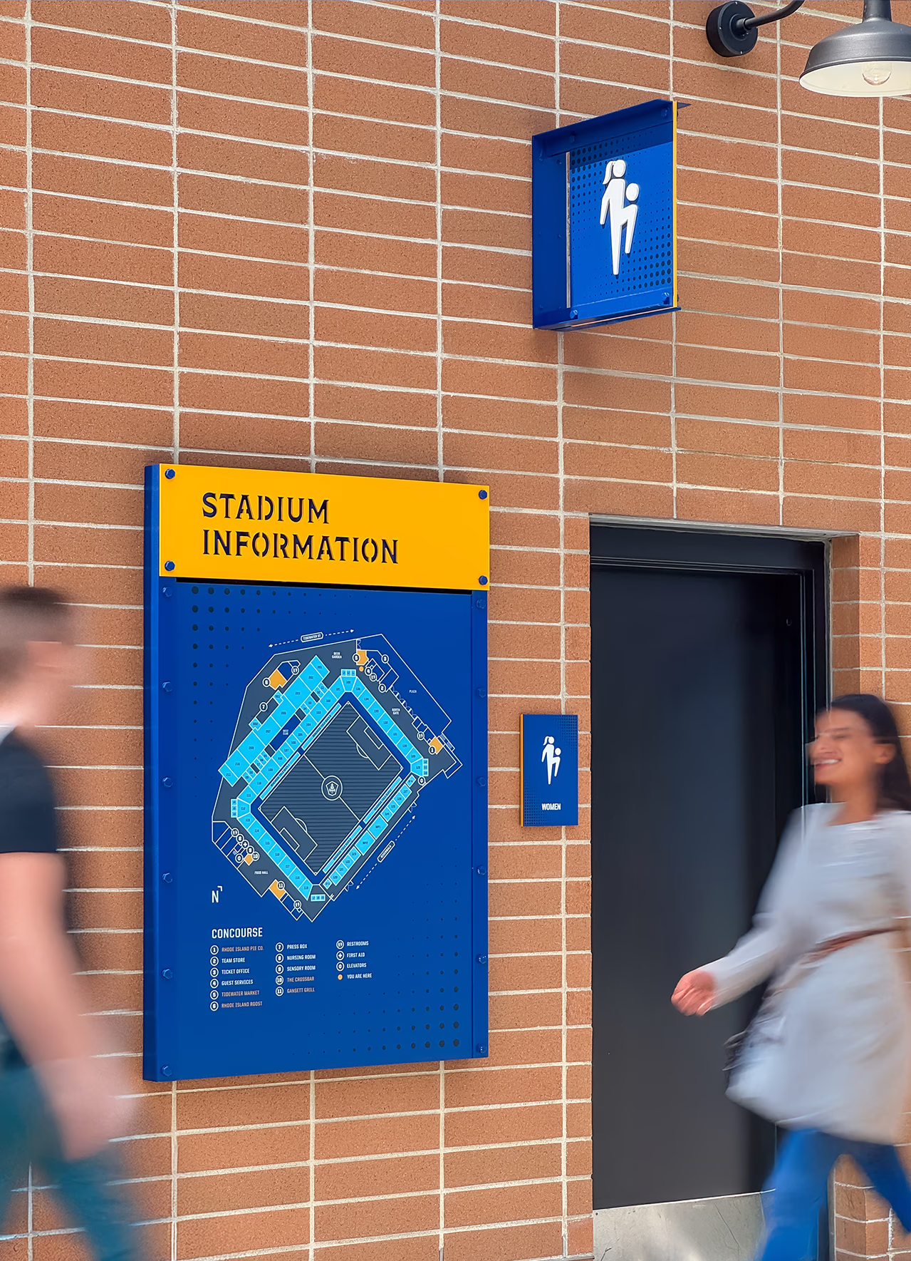 A wall-mounted stadium information map and blue restroom identification signs on brick at Centreville Bank Stadium as fans walk past on the concourse.