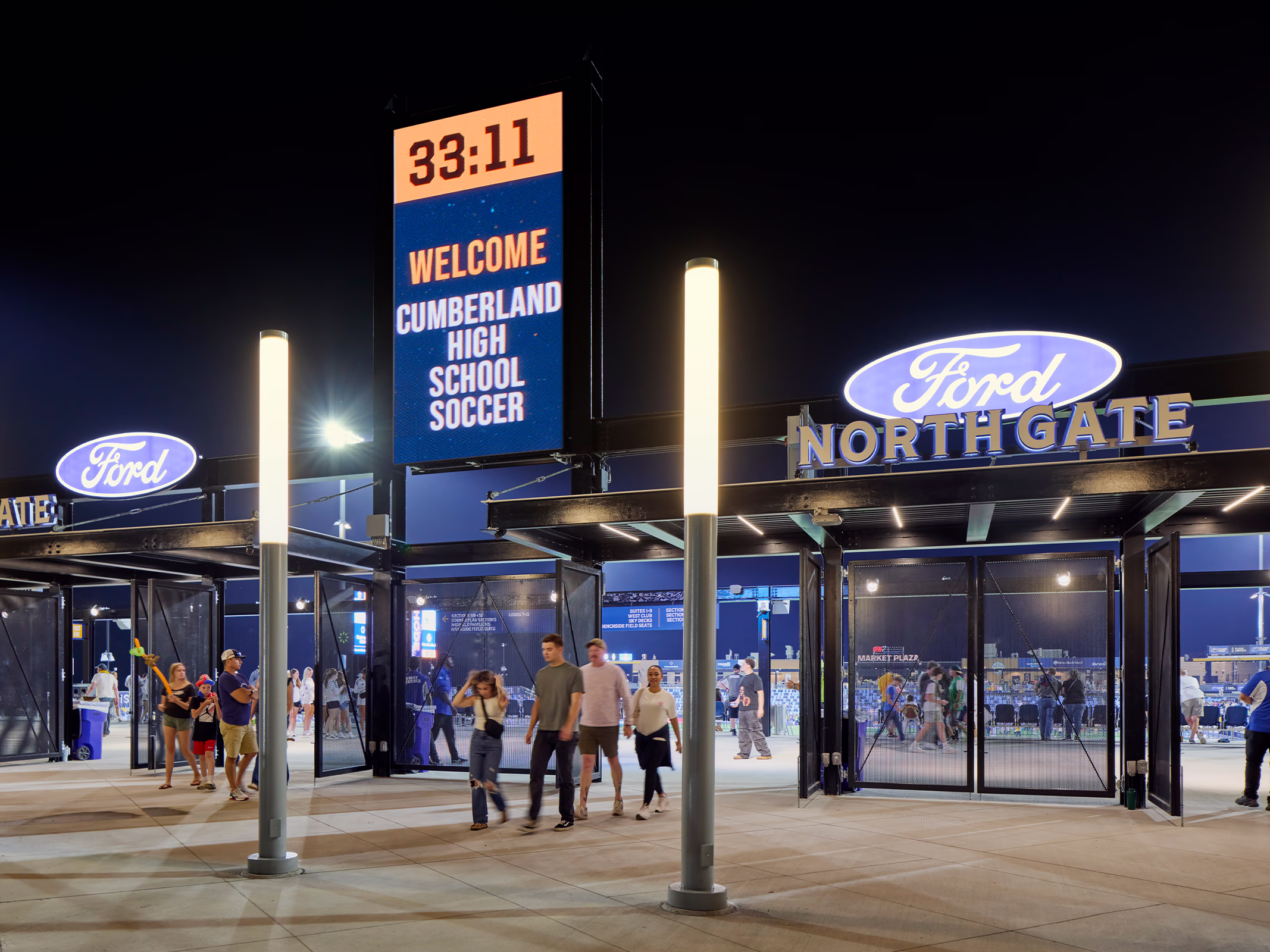 Illuminated Ford North Gate signage and an LED welcome board light the entry plaza at Centreville Bank Stadium as fans walk through at night.