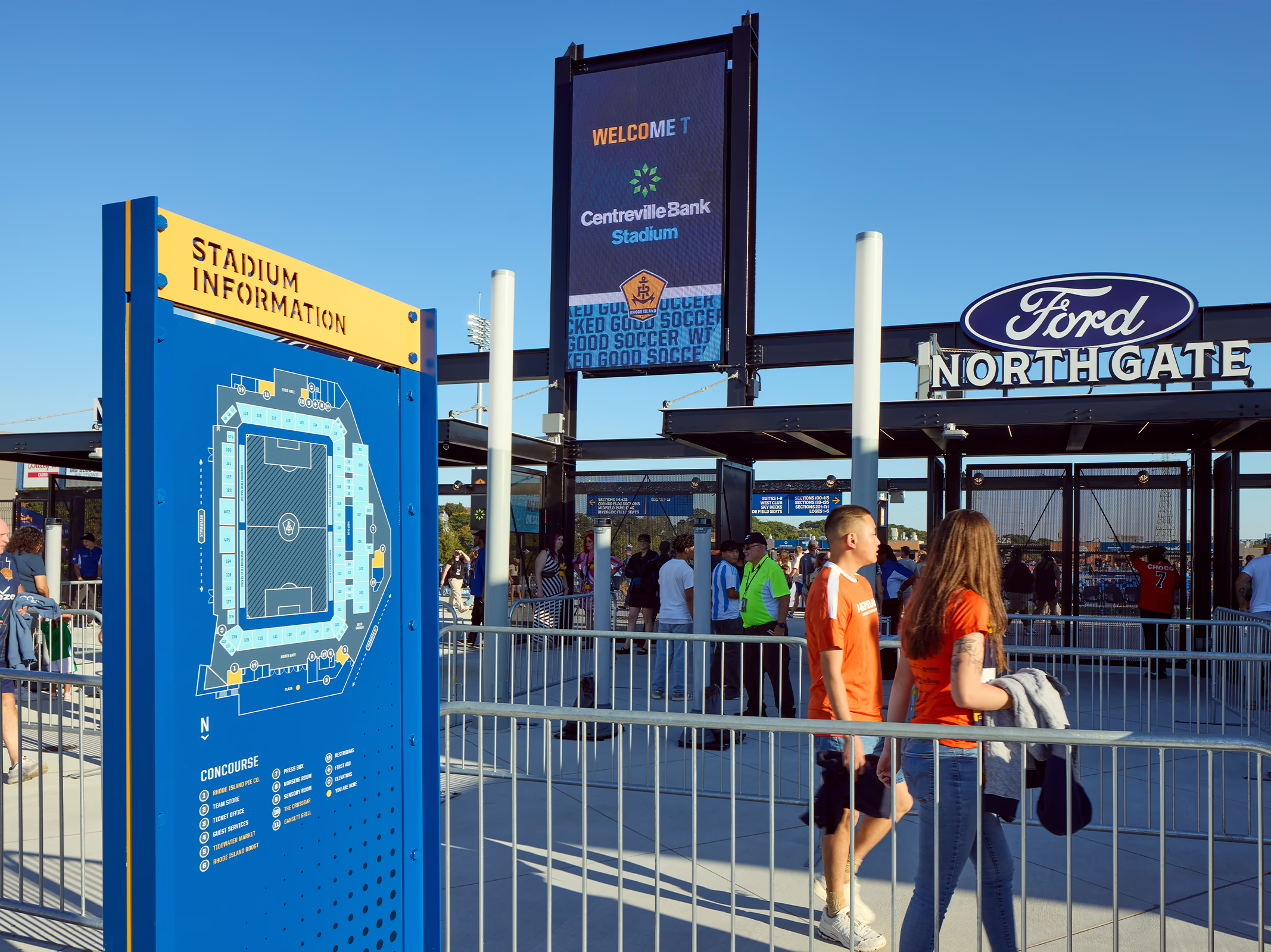 A blue and yellow stadium information kiosk with a concourse map and amenity directory guides fans entering the North Gate plaza at Centreville Bank Stadium.