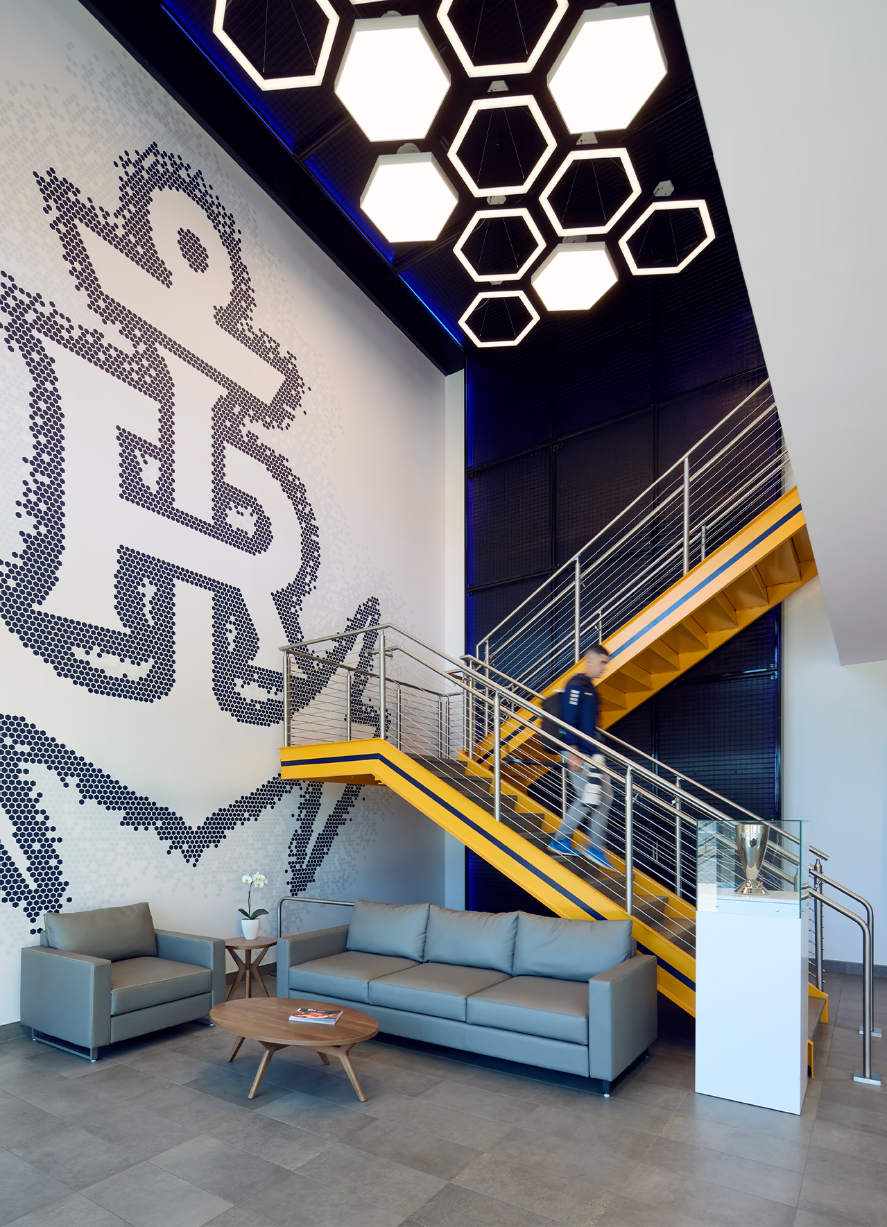 A halftone anchor crest mural rises above the lobby lounge at Centreville Bank Stadium, beside a yellow feature stair and hexagonal pendant lights.