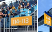 Stadium seating filled with fans and a section number sign in the foreground; paired with an overhead identification sign displaying seat numbers for the section.