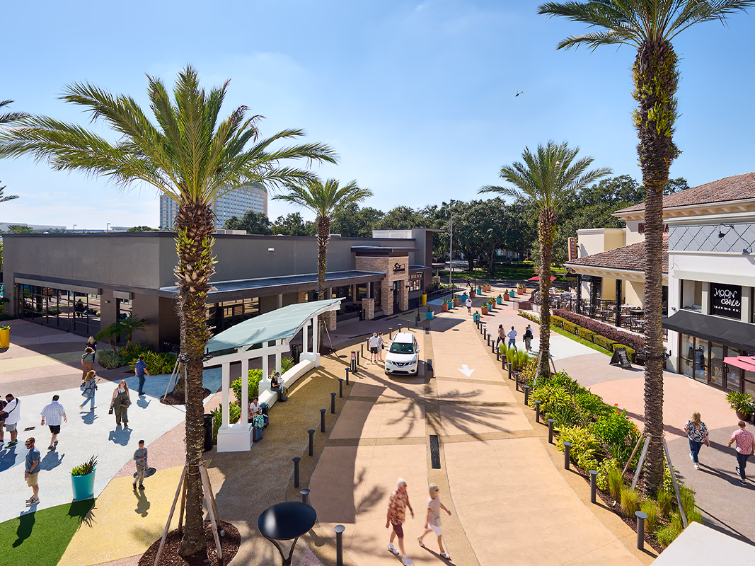 Aerial daytime view of Pointe Orlando's open-air walkway lined with tall palm trees, landscaped greenery, bollards, and retail storefronts, with visitors strolling through the complex under a clear blue sky.
